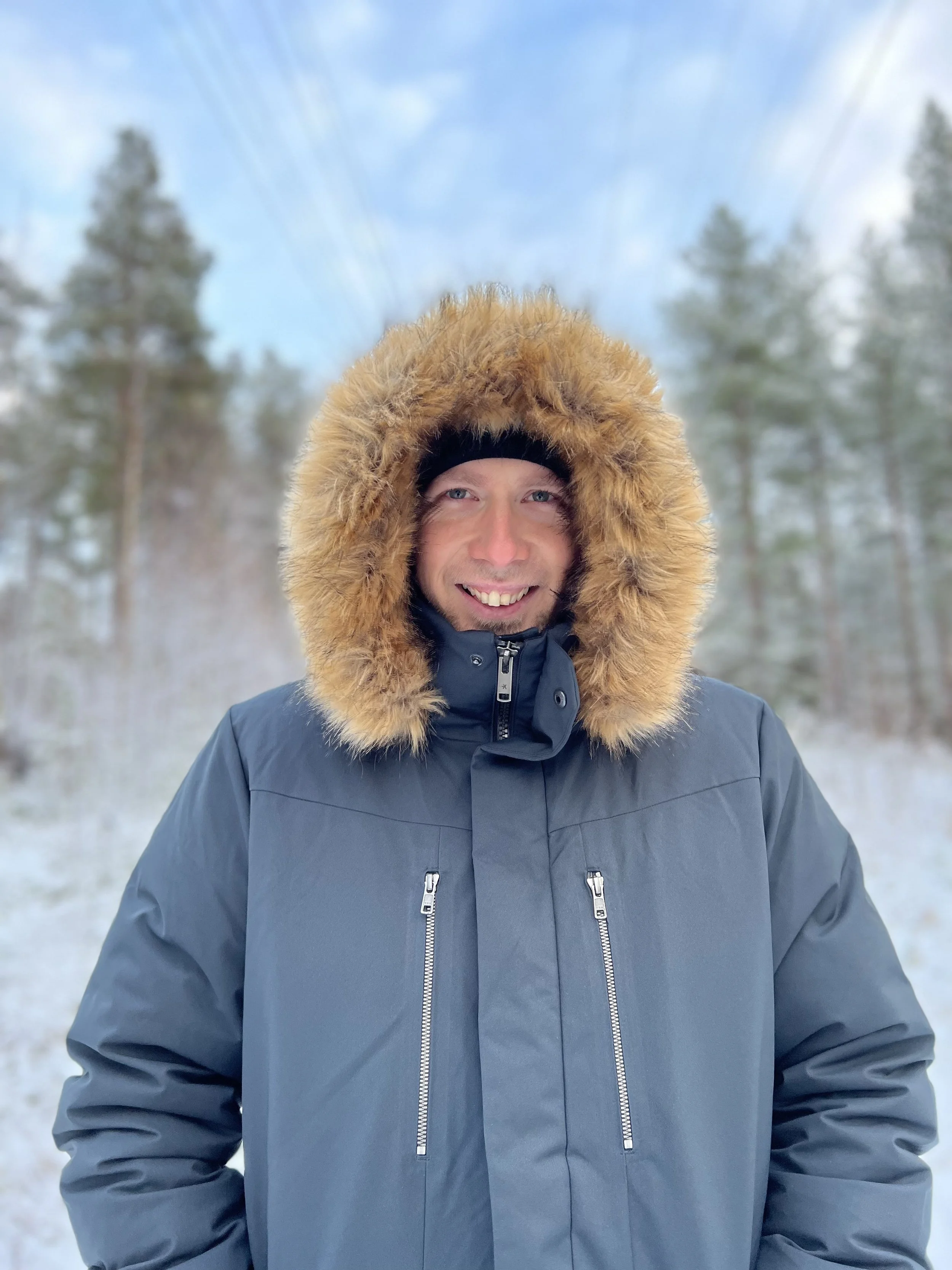 Ein Mann trägt eine dunkelblaue Winterjacke mit Kunstpelzkapuze vor verschneitem Wald, lächelt und schaut in die Kamera.