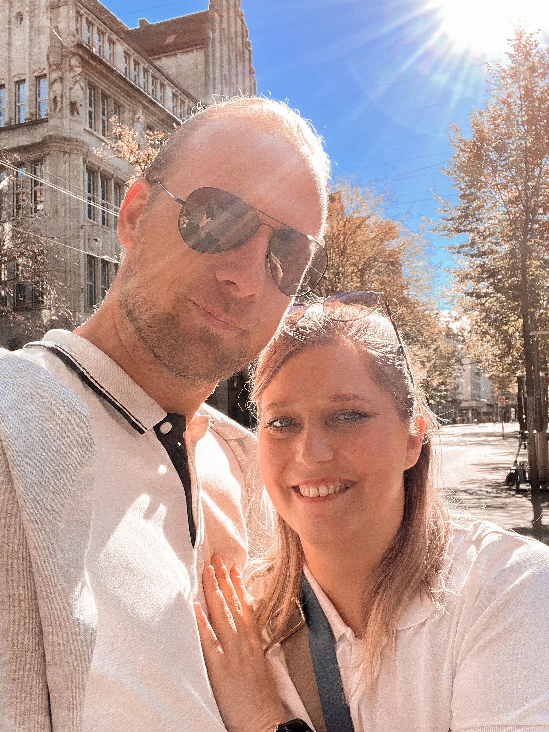 Zwei Menschen, ein Mann mit Sonnenbrille und eine Frau, nehmen ein Selfie auf einer sonnigen Straße auf.