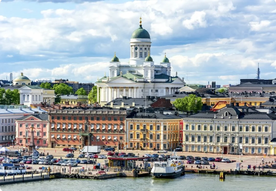 Stadtansicht mit Hafen, mehrstöckigen Gebäuden und einer großen weißen Kirche mit grünen Kuppeln im Hintergrund.