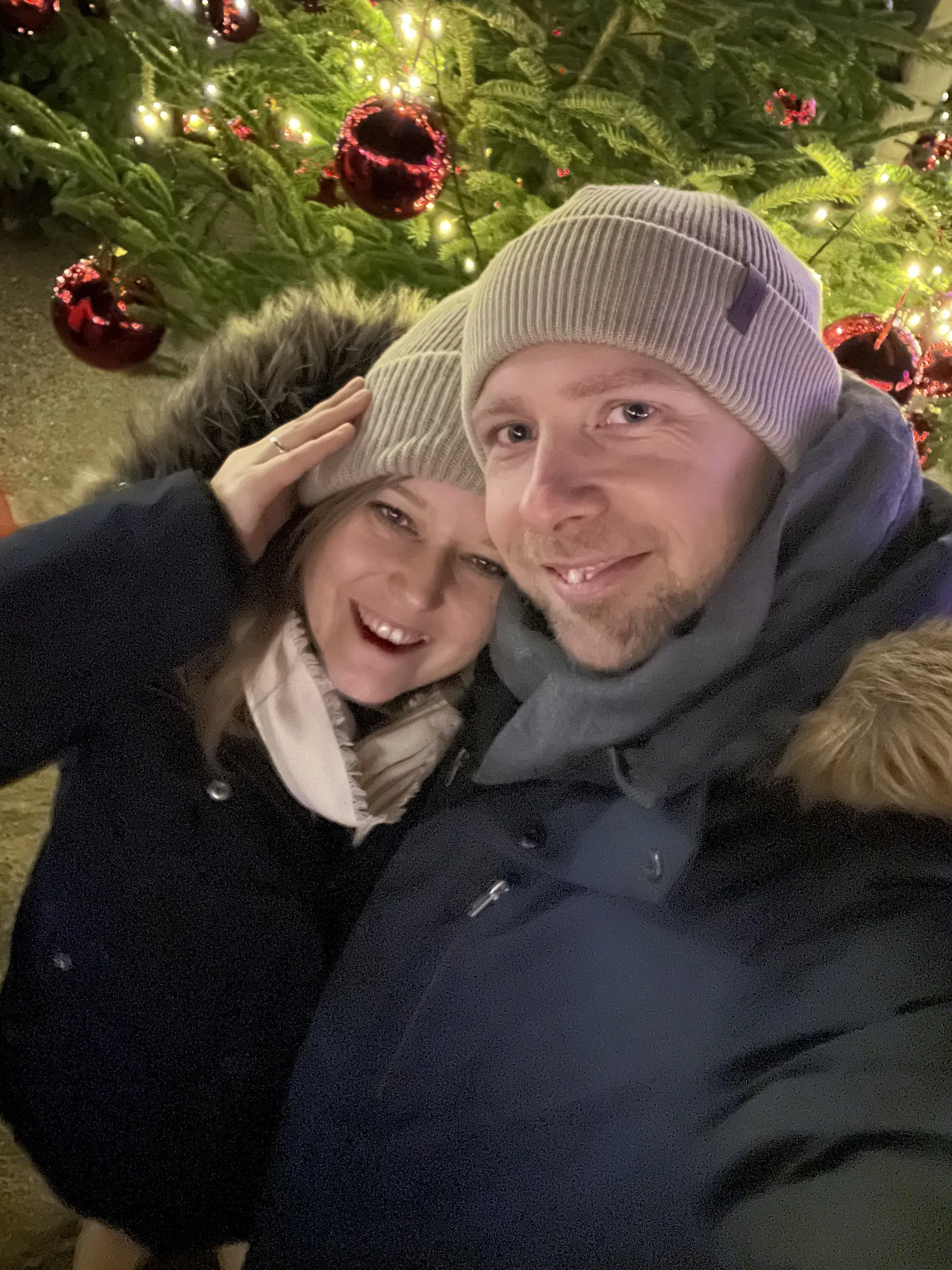 Ein Paar macht ein Selfie vor einem Weihnachtsbaum mit roten Ornamenten und Lichterketten, beide tragen Mützen bei Nacht.