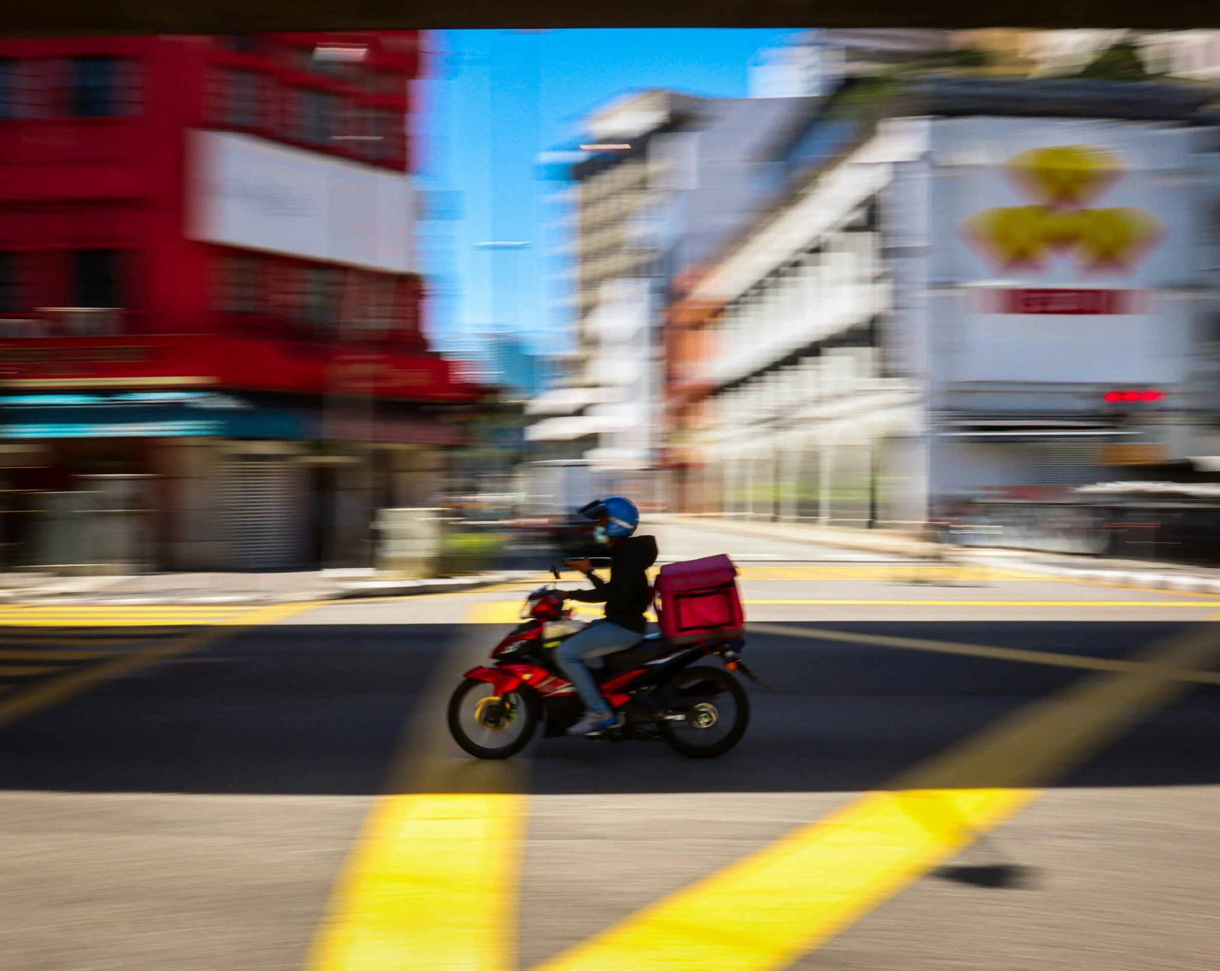 Blurred city street scene with a person riding a motorcycle, colorful buildings, a poster, and yellow lines on the road.