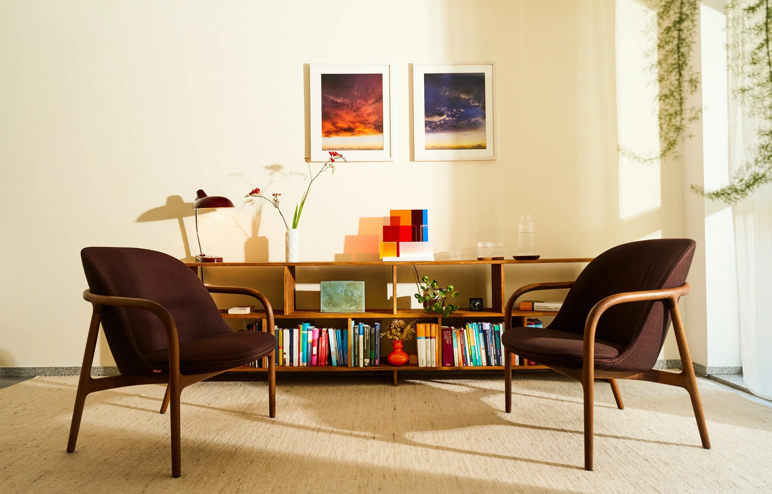 Therapieraum der Privatpraxis Anna Anft in Köln-Sülz mit zwei dunkelbraunen Sesseln, Holzregal mit Büchern und Pflanzen sowie einer Lampe und einem beige Teppich, an einer hellen Wand mit zwei Fotografien von Sonnenuntergängen.
