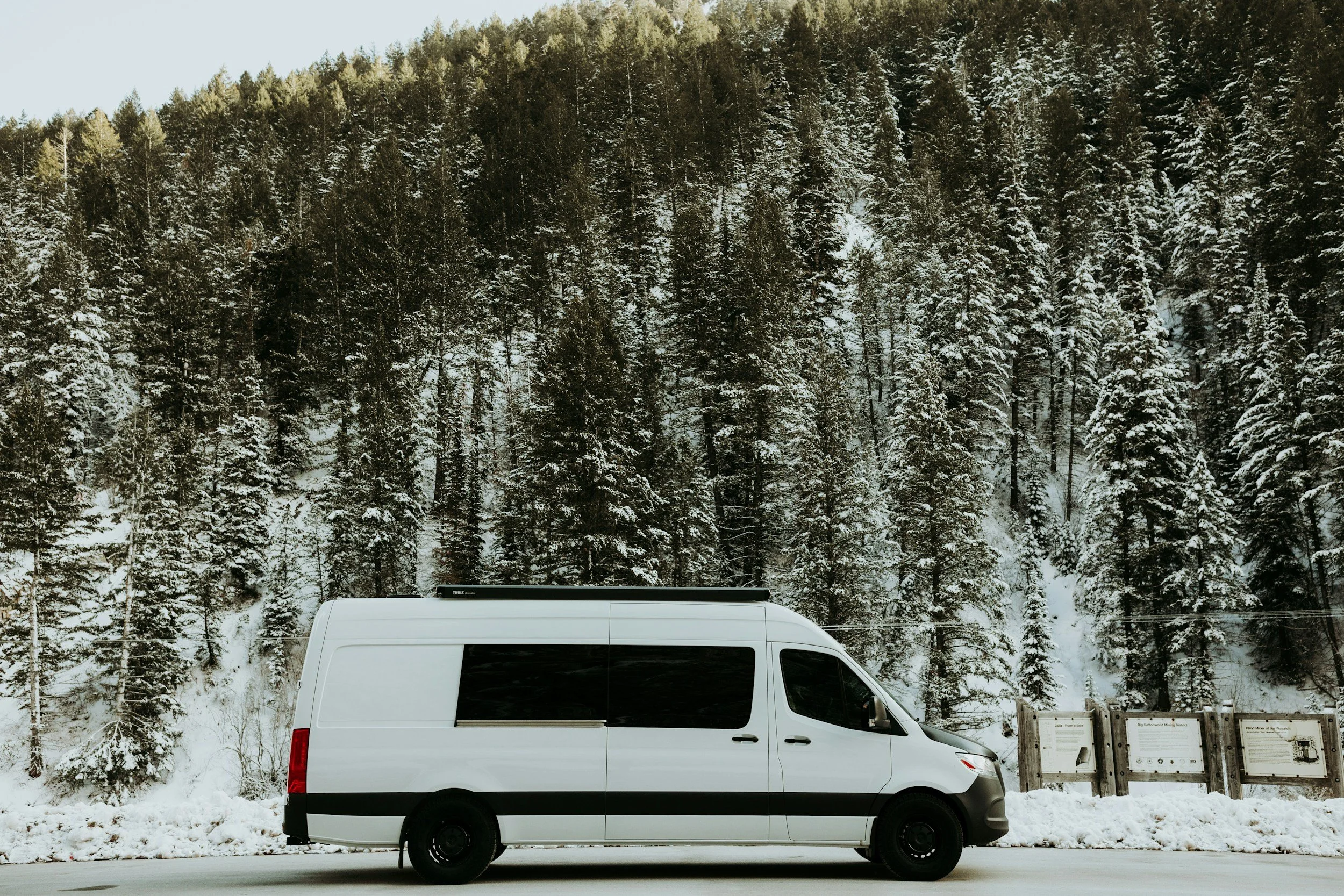 White van parked in front of a snow-covered forest with evergreen trees and informational signs near the roadside.