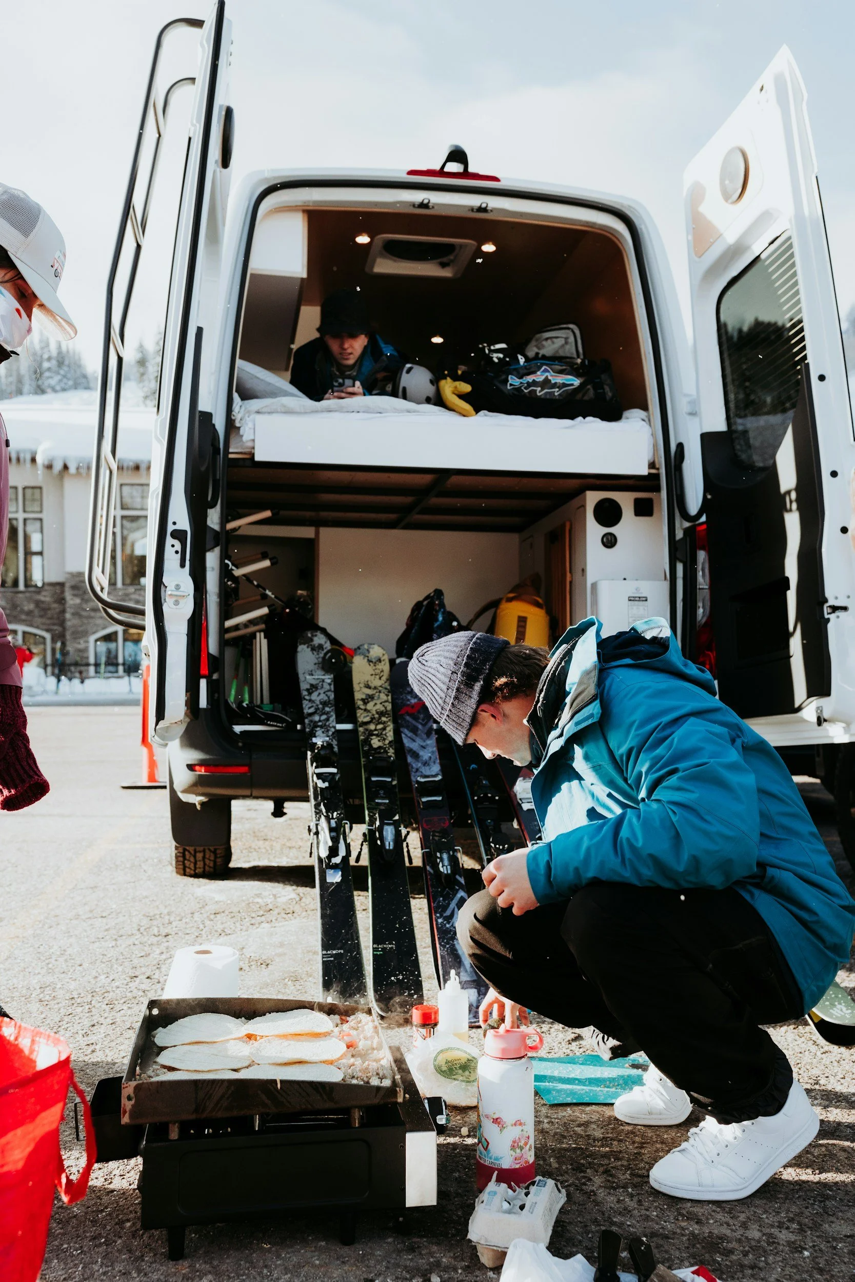 People preparing food outside next to a vehicle with an upper sleeping area, winter clothing, skis, and snow on the ground.