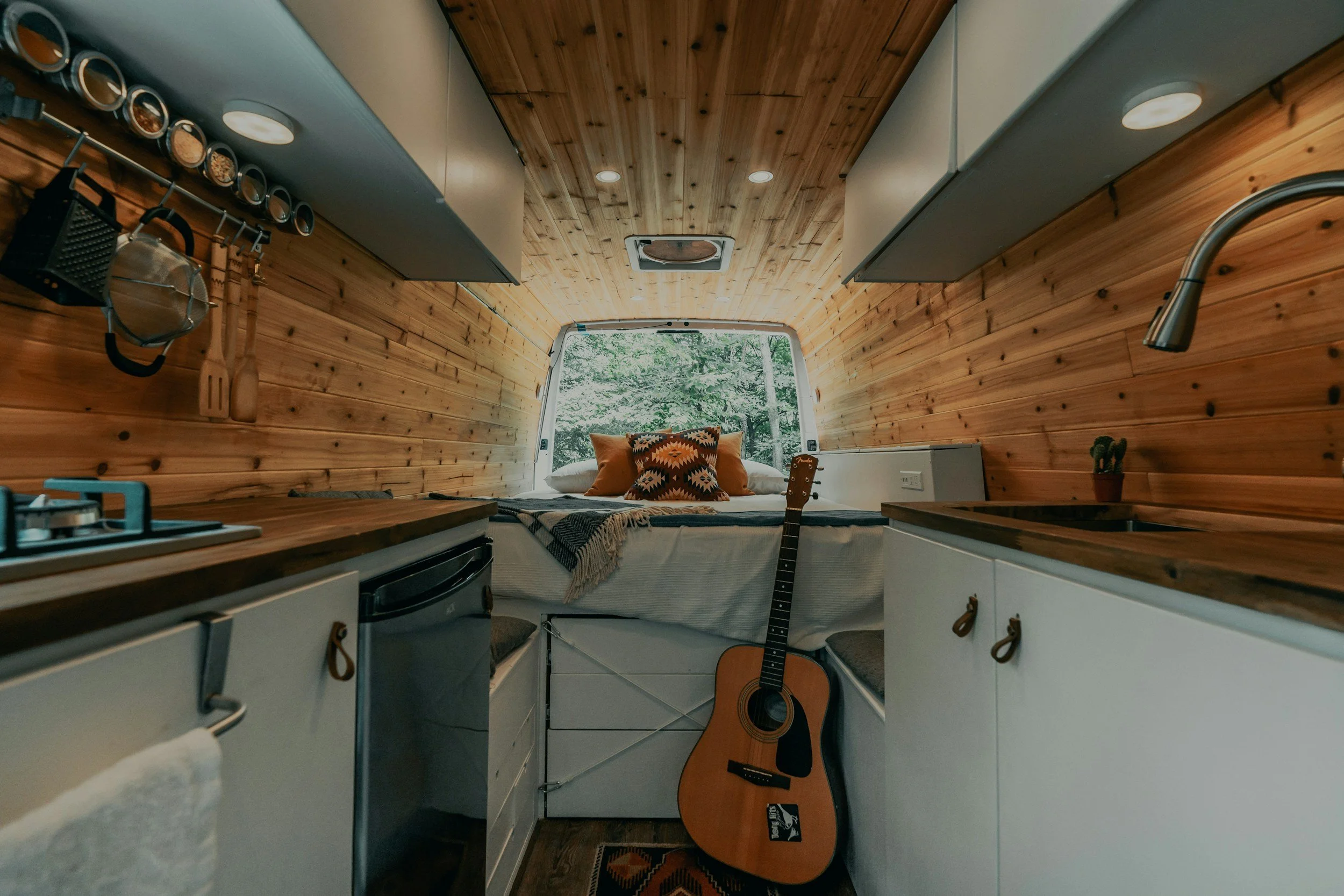 Interior of a tiny wooden camper with a kitchen area and a bed at the far end near a window with trees outside. A guitar is leaning against the bed, and there are pots and kitchen utensils hanging on one side.  Interior of camper van.
