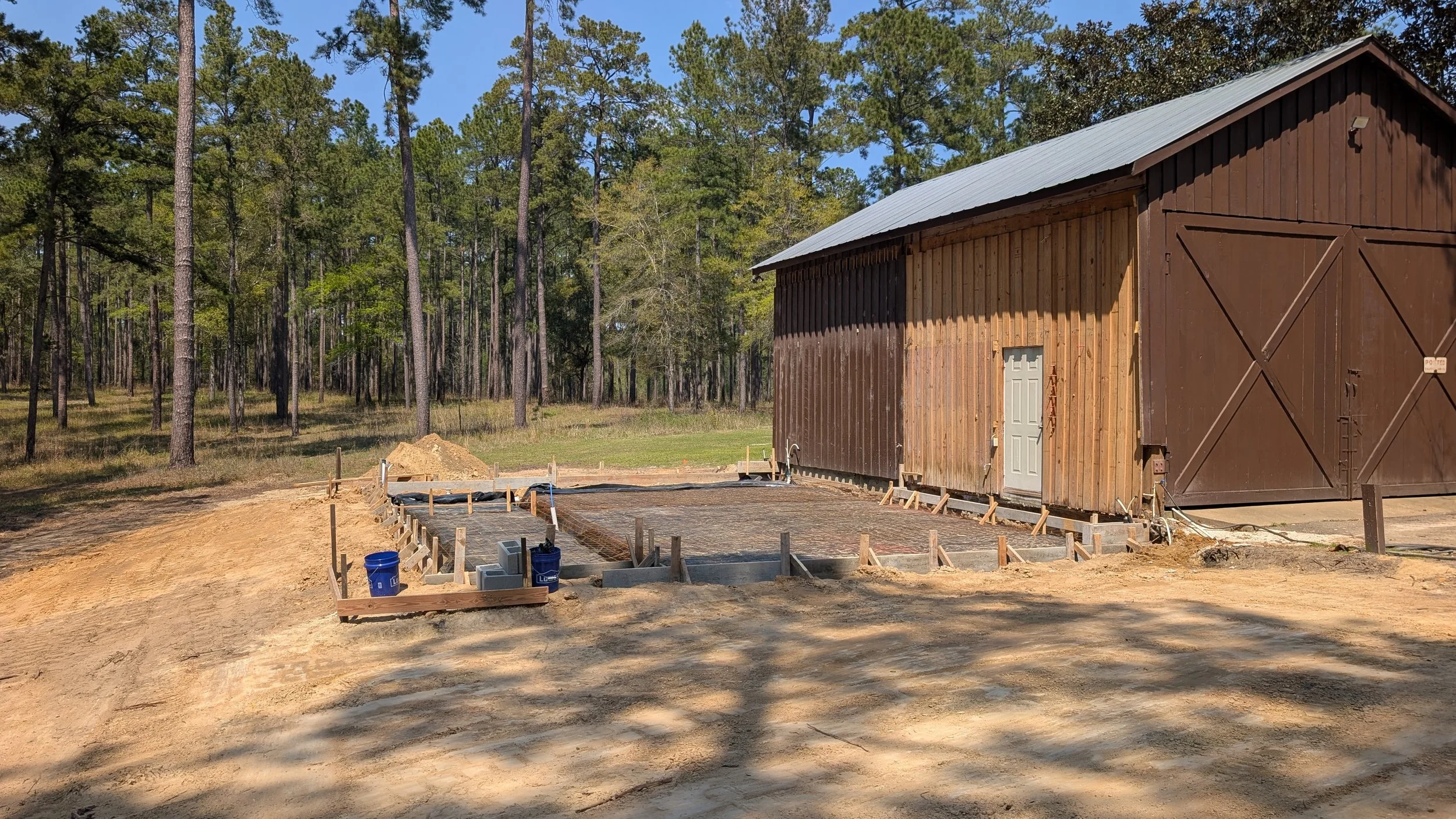 Construction site with a wooden barn and concrete foundation being prepared in a wooded area.