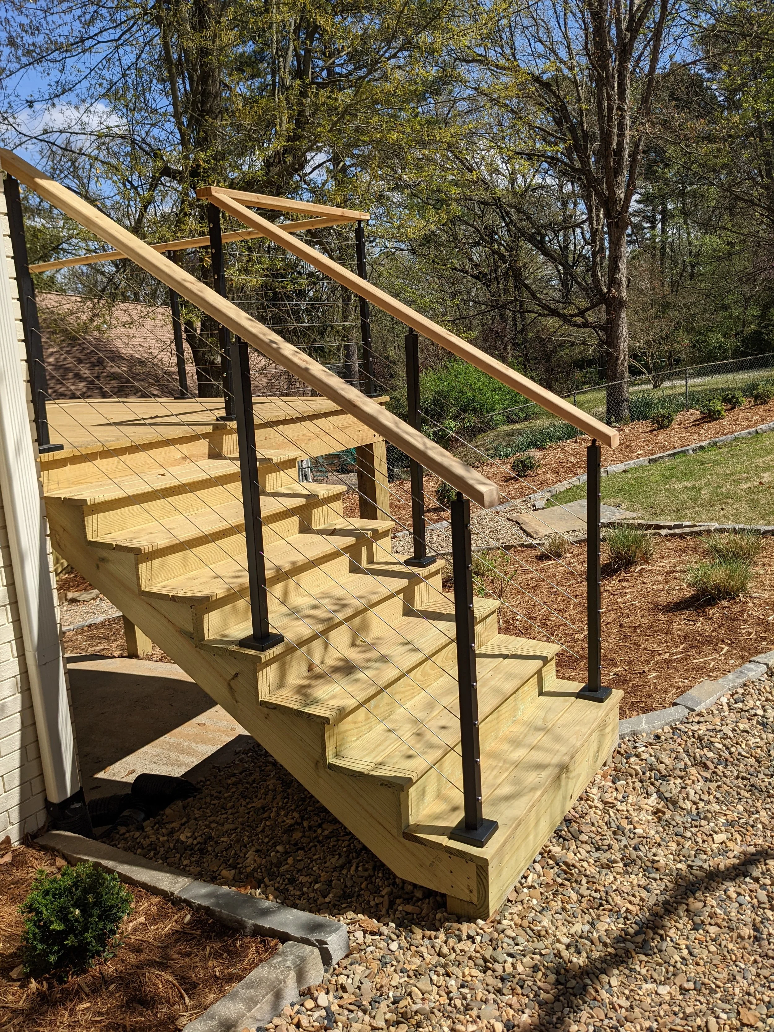 Newly constructed outdoor wooden staircase with metal cable railing next to a house, leading down to a landscaped yard with gravel and a small shrub, in a backyard with trees and clear blue sky.