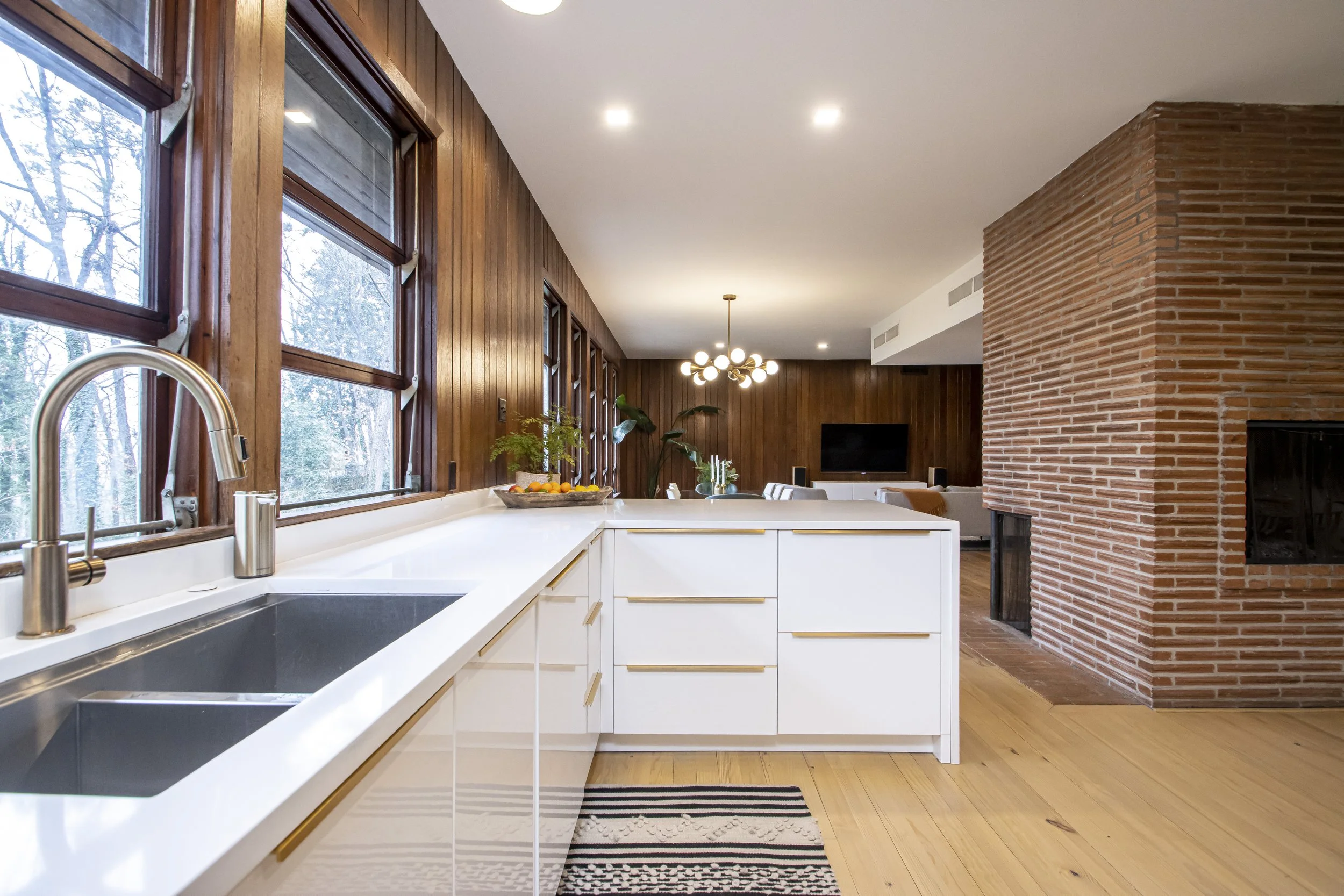 Modern kitchen with white cabinets, a double sink, large windows, a fruit bowl, wood paneling on walls, and a brick fireplace in the open living room area.