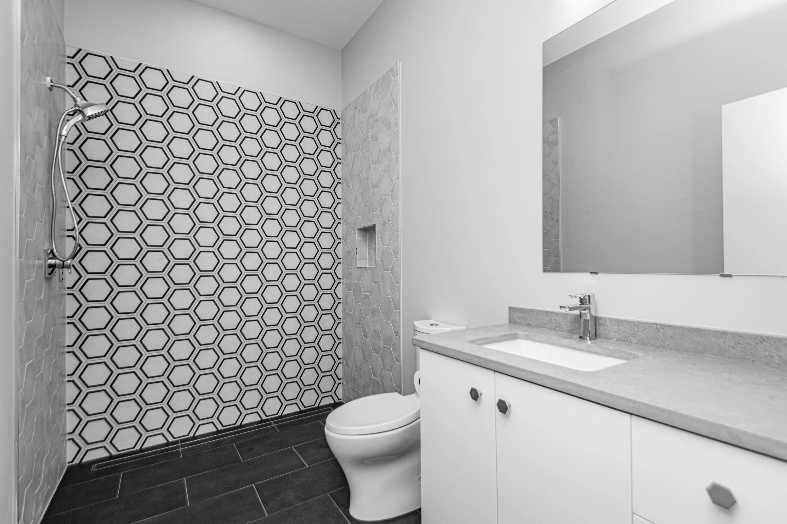 Modern bathroom with a shower area featuring black and white geometric tiles, white vanity with a round vessel sink, large mirror, and white toilet.