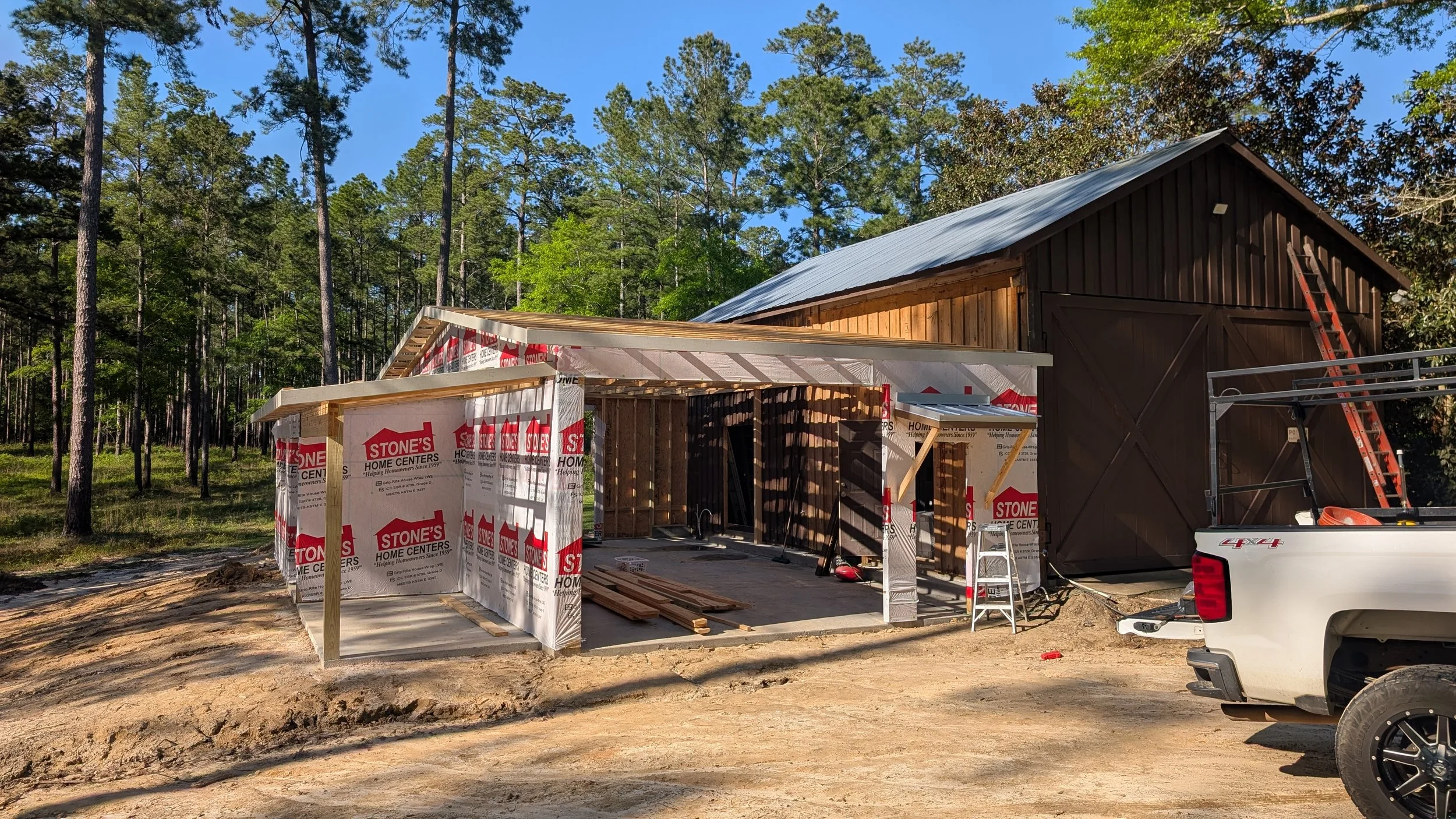 Construction site with a house being built, surrounded by trees, with partial framing and a completed section with dark wood siding, ladder, scaffolding, and a pickup truck.