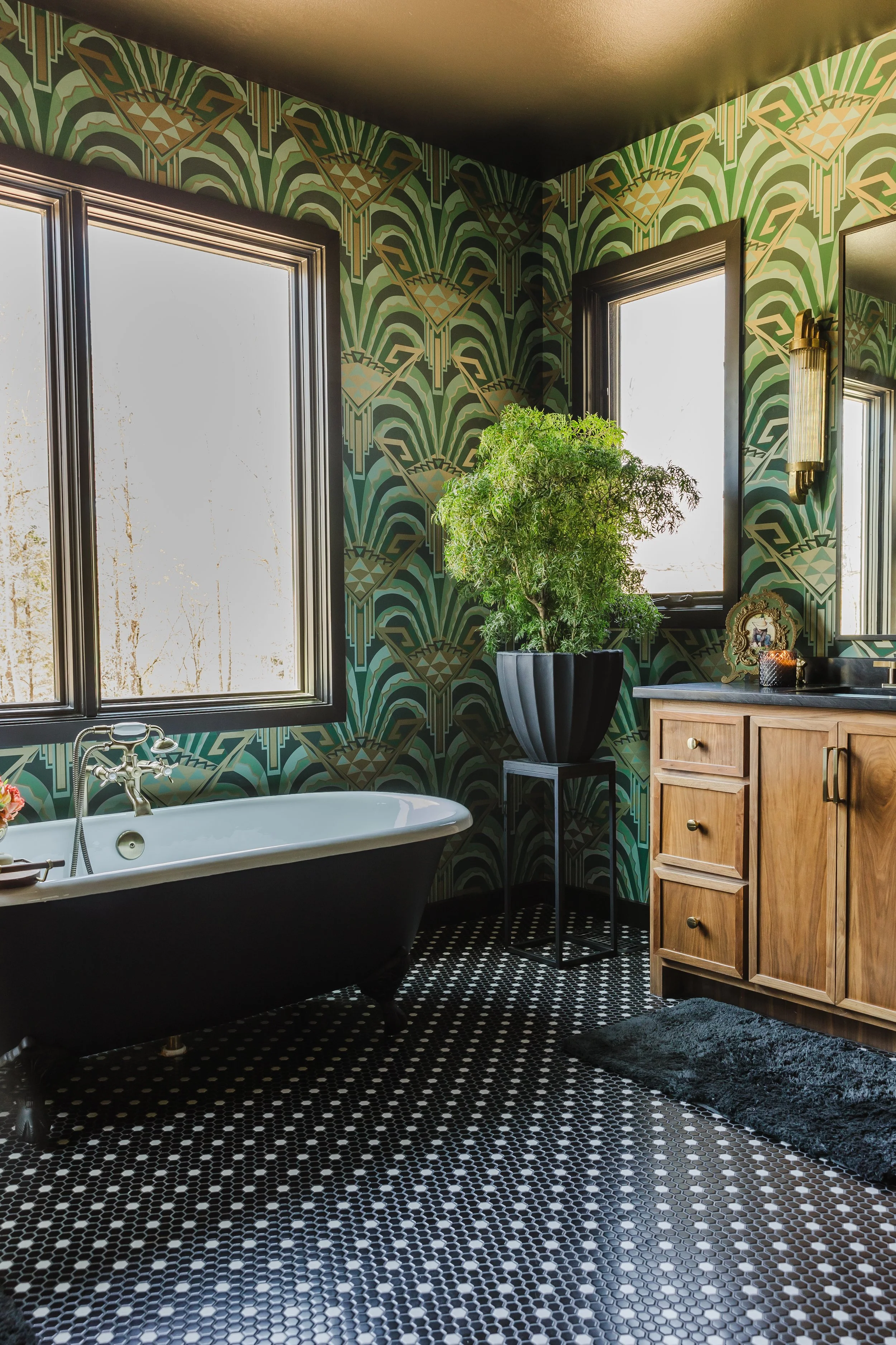 A bathroom with black and white hexagonal floor tiles, a clawfoot bathtub, a wooden vanity with drawers, a large potted plant on a black stand, patterned green wallpaper, and two windows letting in light.