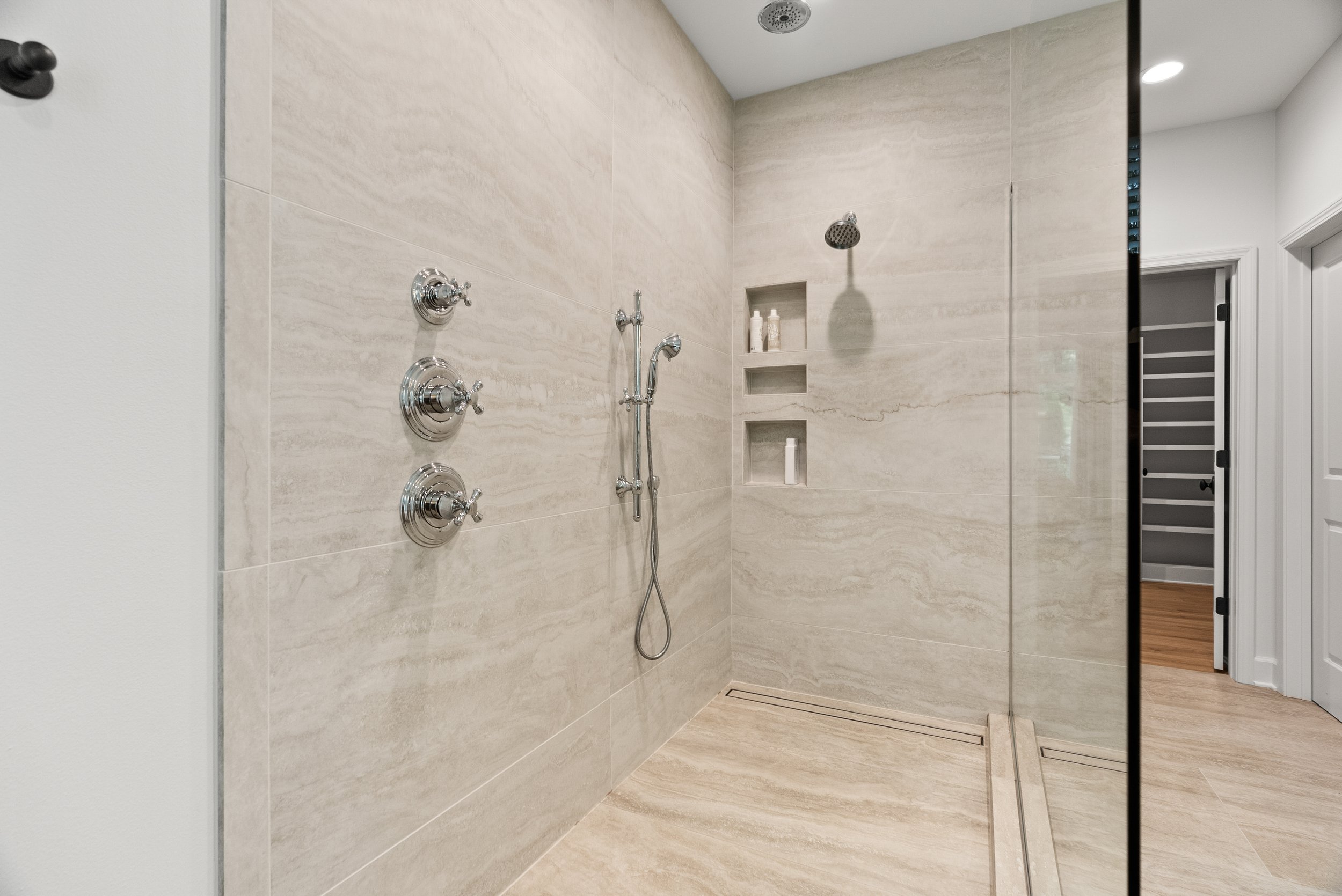 A modern shower with beige marble tiles, a rainfall showerhead, a handheld shower on a sliding bar, three water control knobs, and built-in wall niches with toiletries.