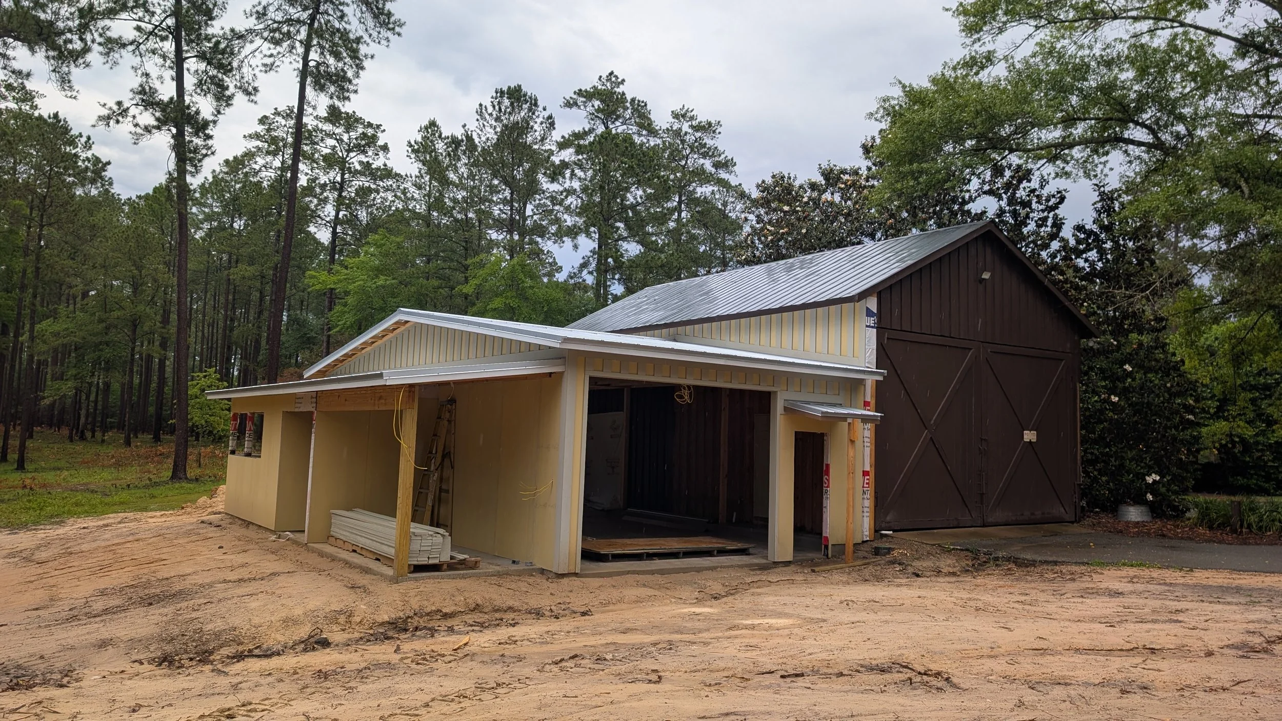 Construction site with a partially built small building with a yellow exterior and a metal roof, next to a larger dark barn, surrounded by a wooded area with tall trees and cloudy sky.