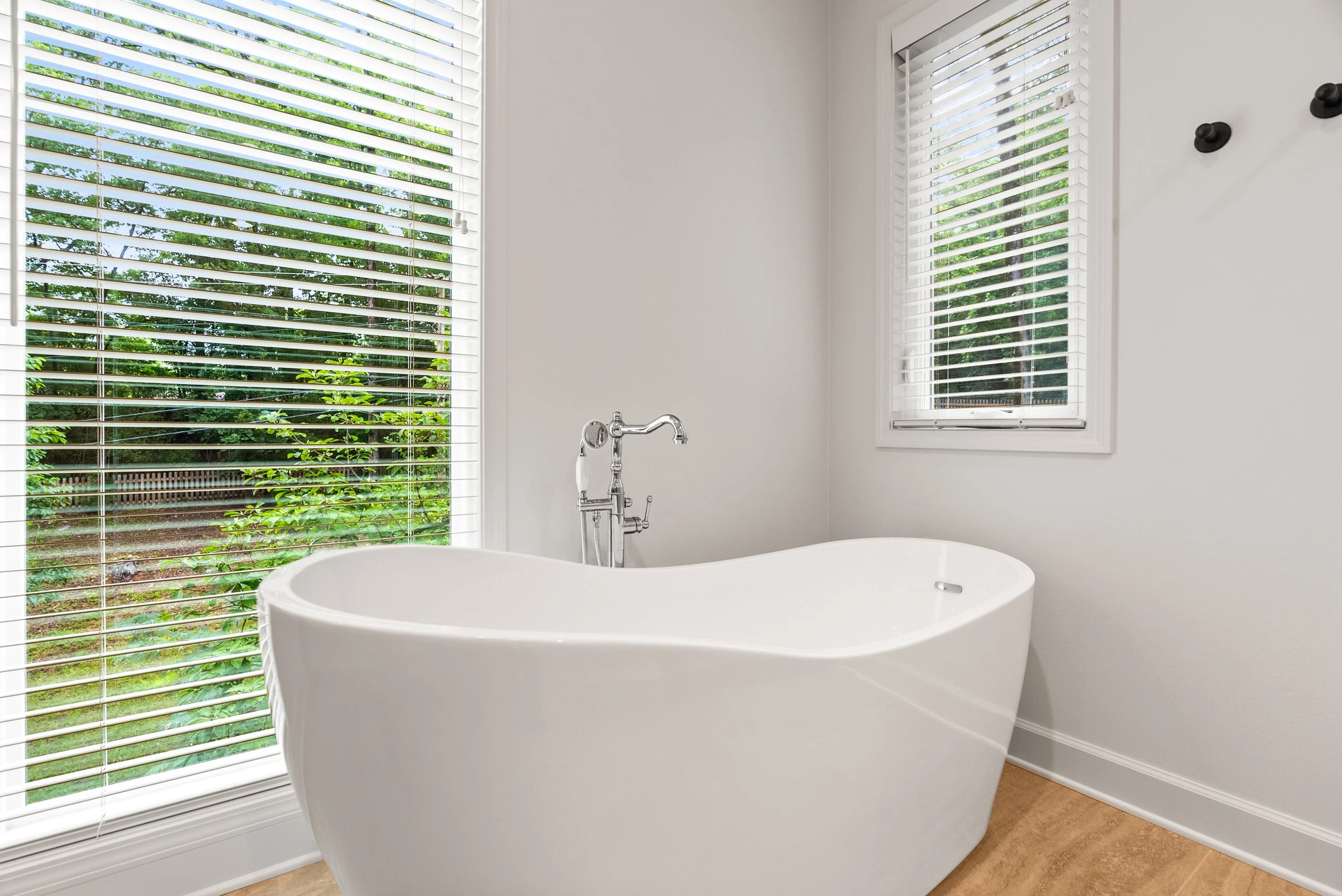 A white freestanding bathtub in a bathroom with white walls and brown flooring. Two large windows with white blinds showing greenery outside.
