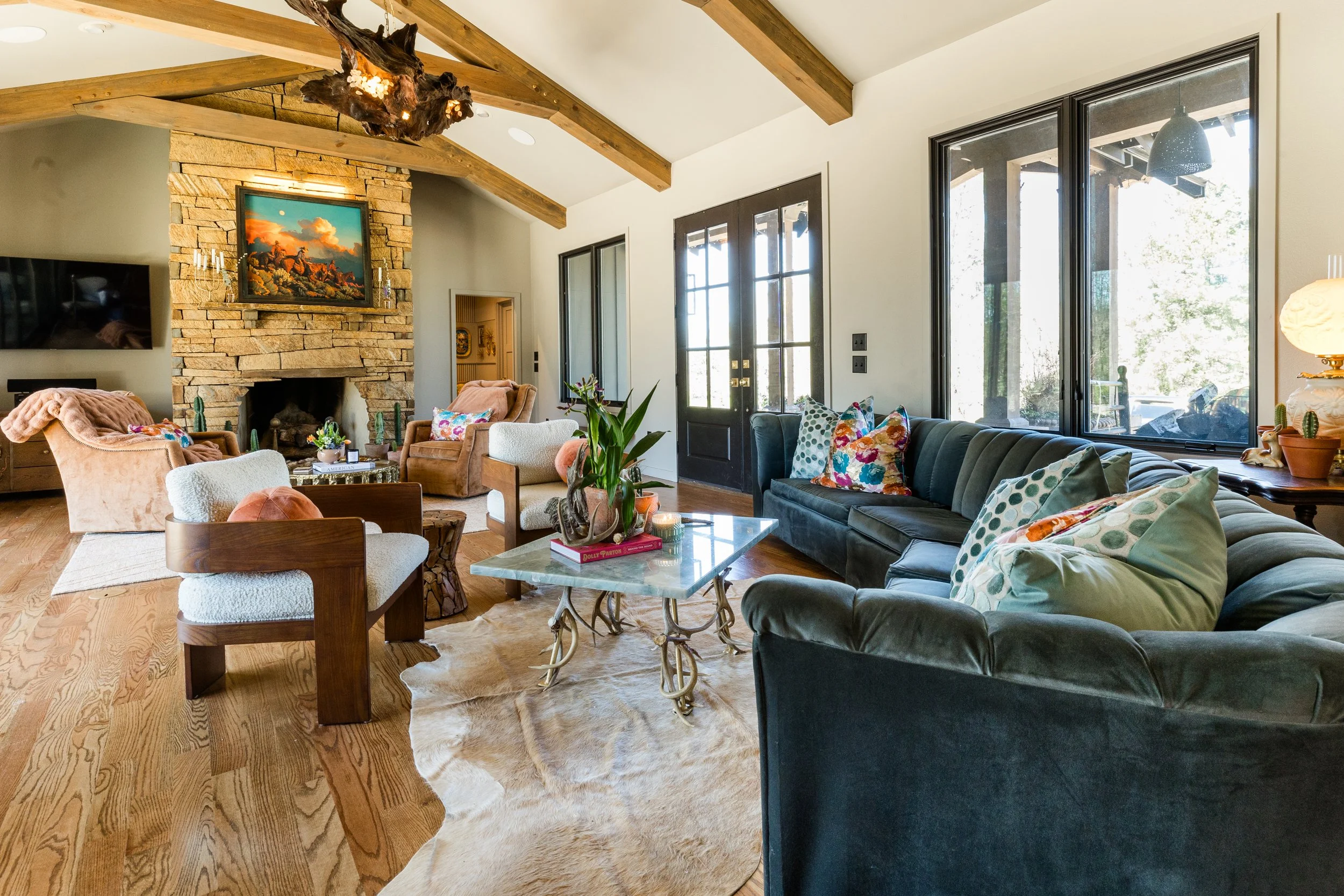 Living room with large windows, a stone fireplace, and a high ceiling with exposed wooden beams. The room has various armchairs and a sofa with colorful pillows, a coffee table with decorative items, and a cowhide rug on the wooden floor.