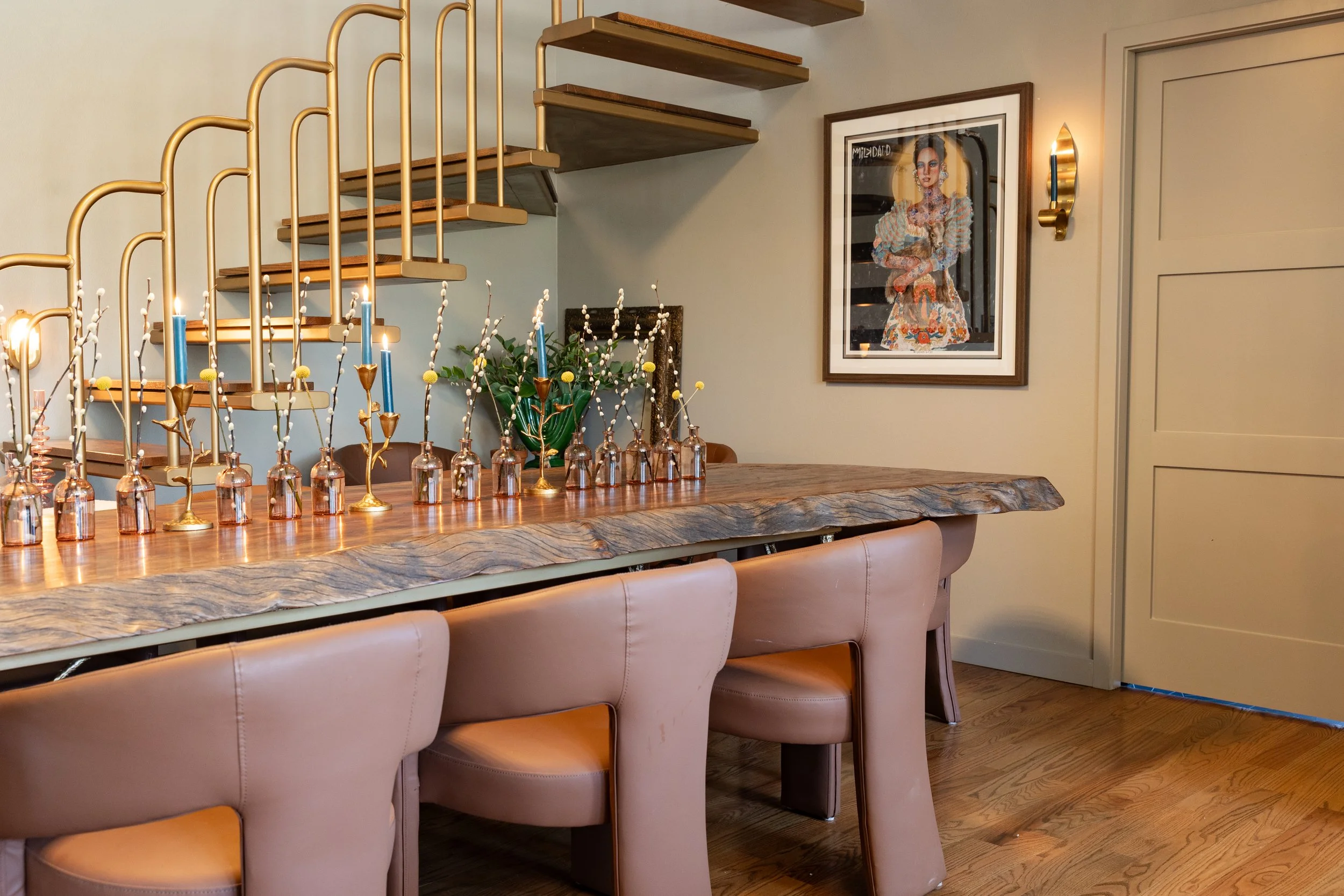 A dining space with a wooden table and beige upholstered chairs. A gold candle holder with blue candles and decorative branches in glass bottles are on the table. There are floating wooden shelves, a large framed artwork of a woman, and a gold wall s
