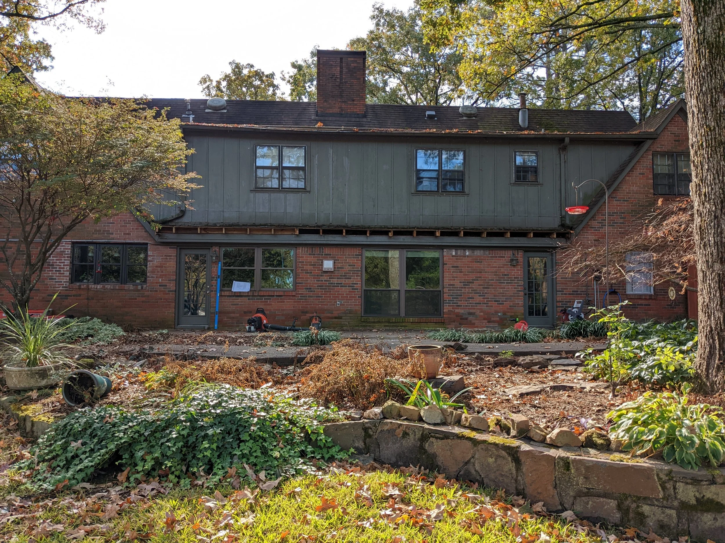 Rear view of a two-story brick house with an uneven gutter system and trees with autumn foliage in the background. The yard is under renovation, with tools and construction materials scattered around.