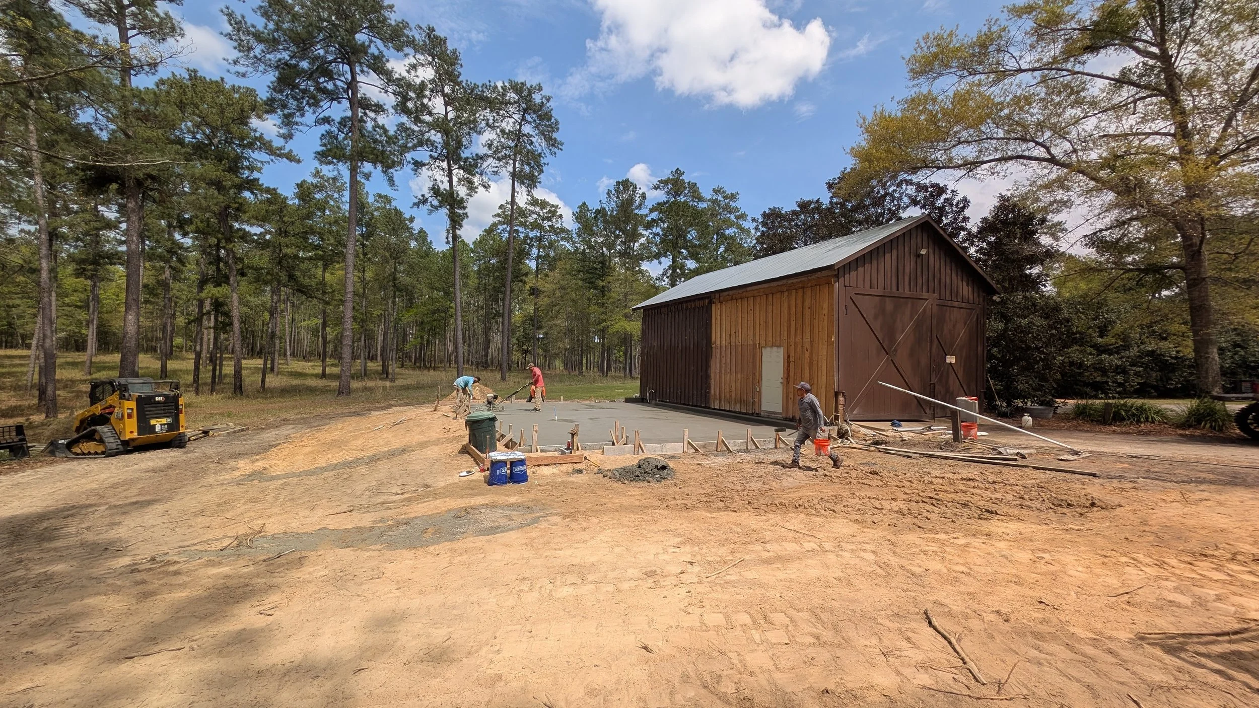 Construction workers pouring concrete in front of a wooden barn in a forested area with tall pine trees and a partly cloudy sky.