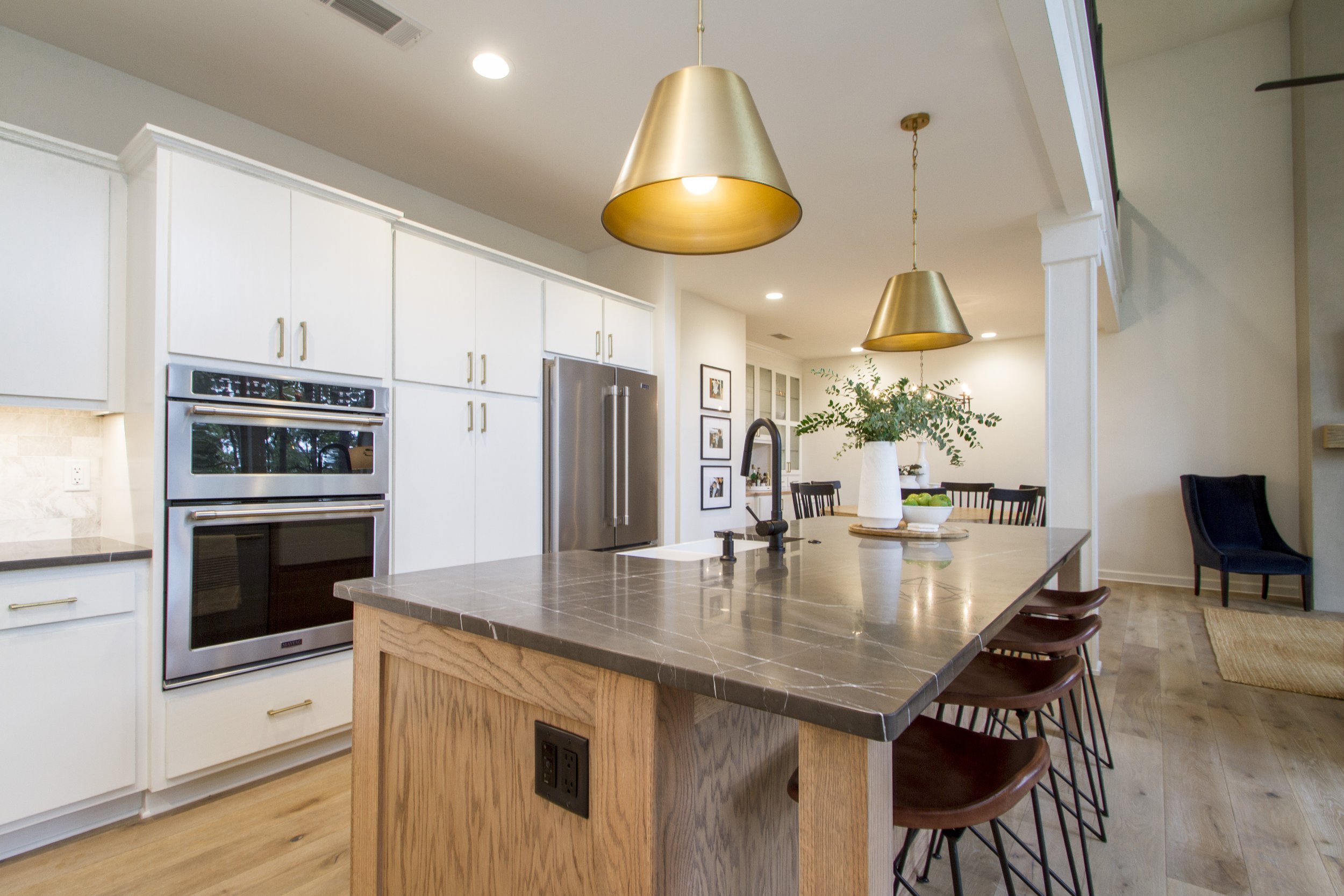 Modern kitchen with white cabinetry, stainless steel refrigerator, black oven, and a kitchen island with a gray tiled countertop. Two gold pendant lights hang above the island. There is a vase with green foliage on the island and dining area in the b