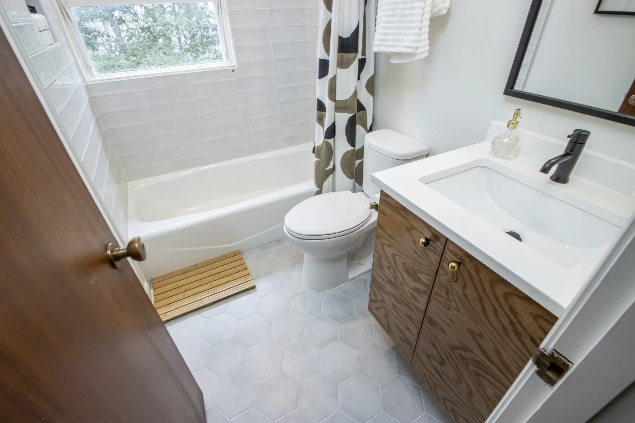 View of a small bathroom with a window, bathtub with shower curtain, toilet, and a wooden vanity with a sink and mirror. The bathroom has white tiled walls and hexagonal floor tiles.
