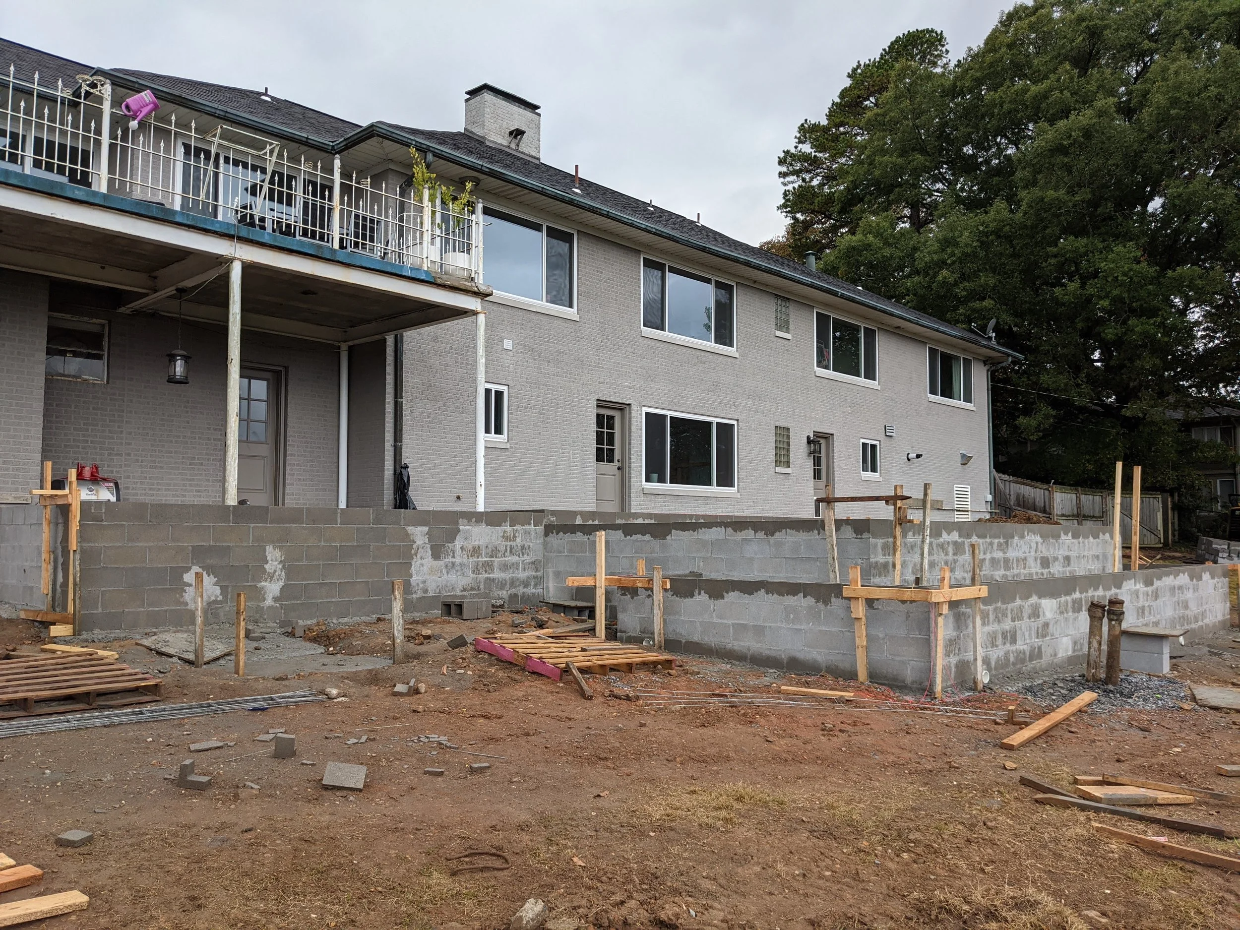 Backyard under construction with cinder block wall, wooden scaffolding, and construction materials, in front of a two-story house with large windows and a balcony.