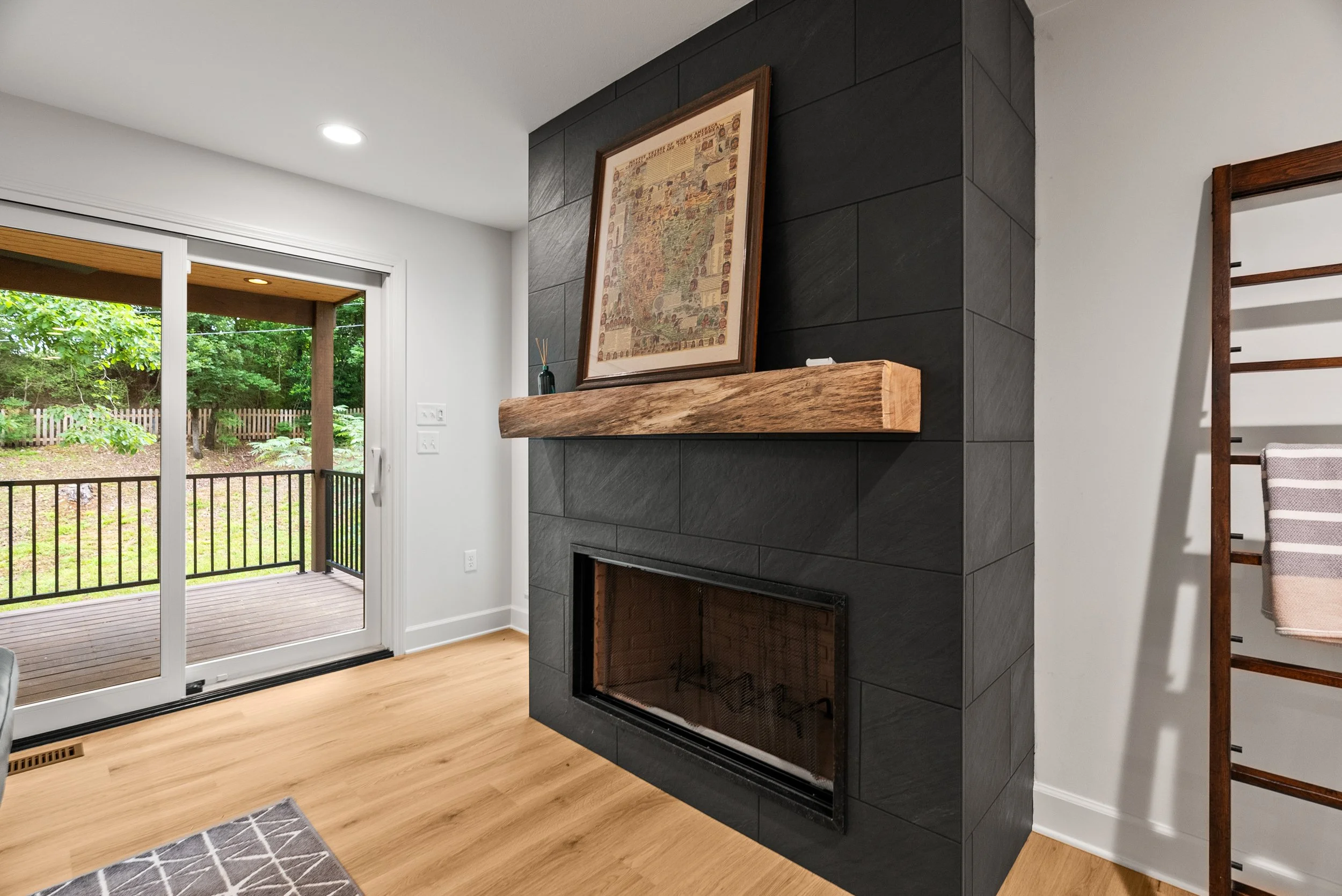 A living room with sliding glass door leading to a wooden porch with green trees outside. Inside, a black tiled fireplace with a wooden mantle and a framed artwork on top. Part of a striped armchair and a geometric-patterned rug are visible.