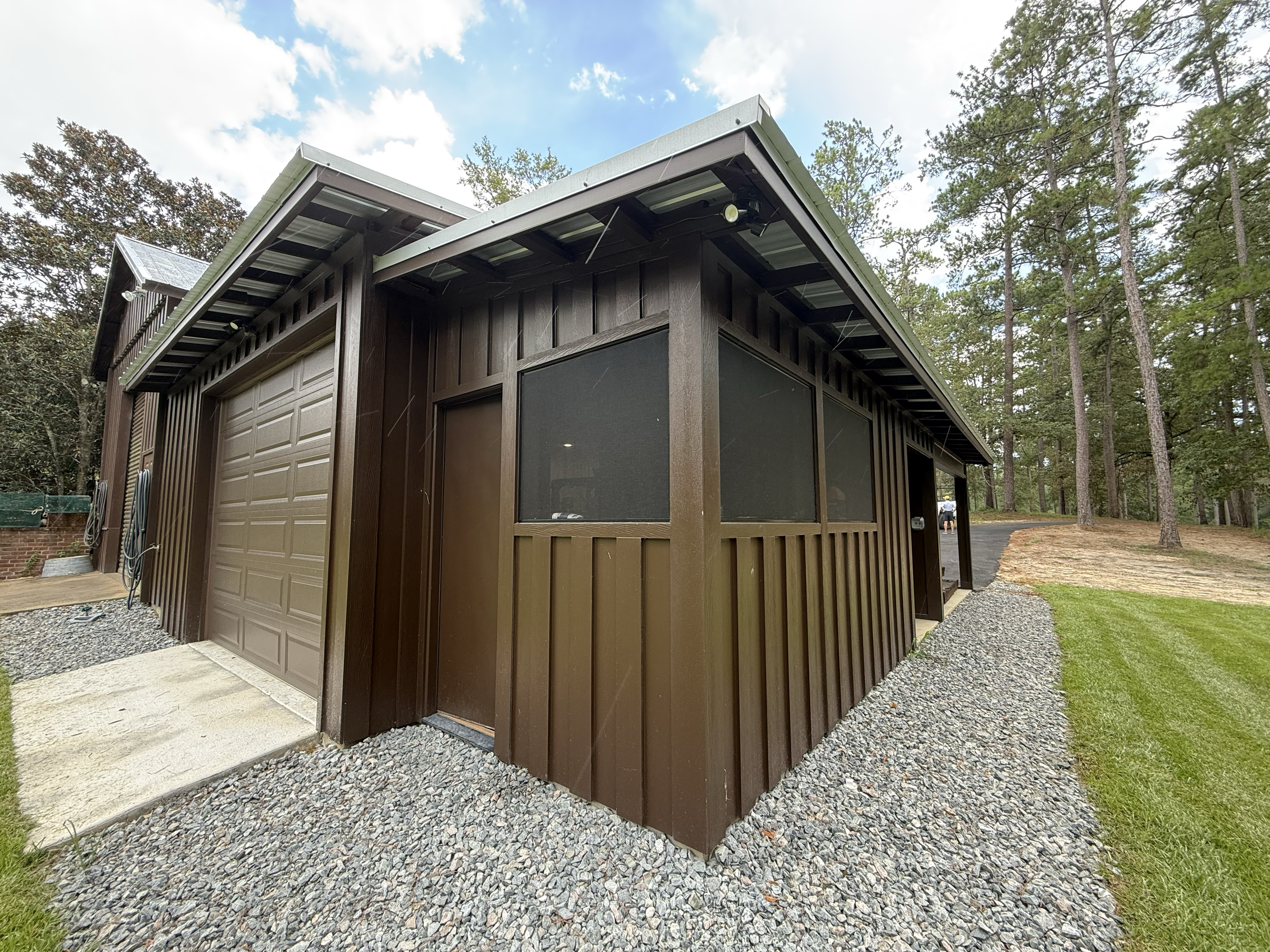 A brown garage with a roll-up door, and a black screen window on the side, surrounded by a gravel yard and a wooded area with tall trees and a partly cloudy sky.