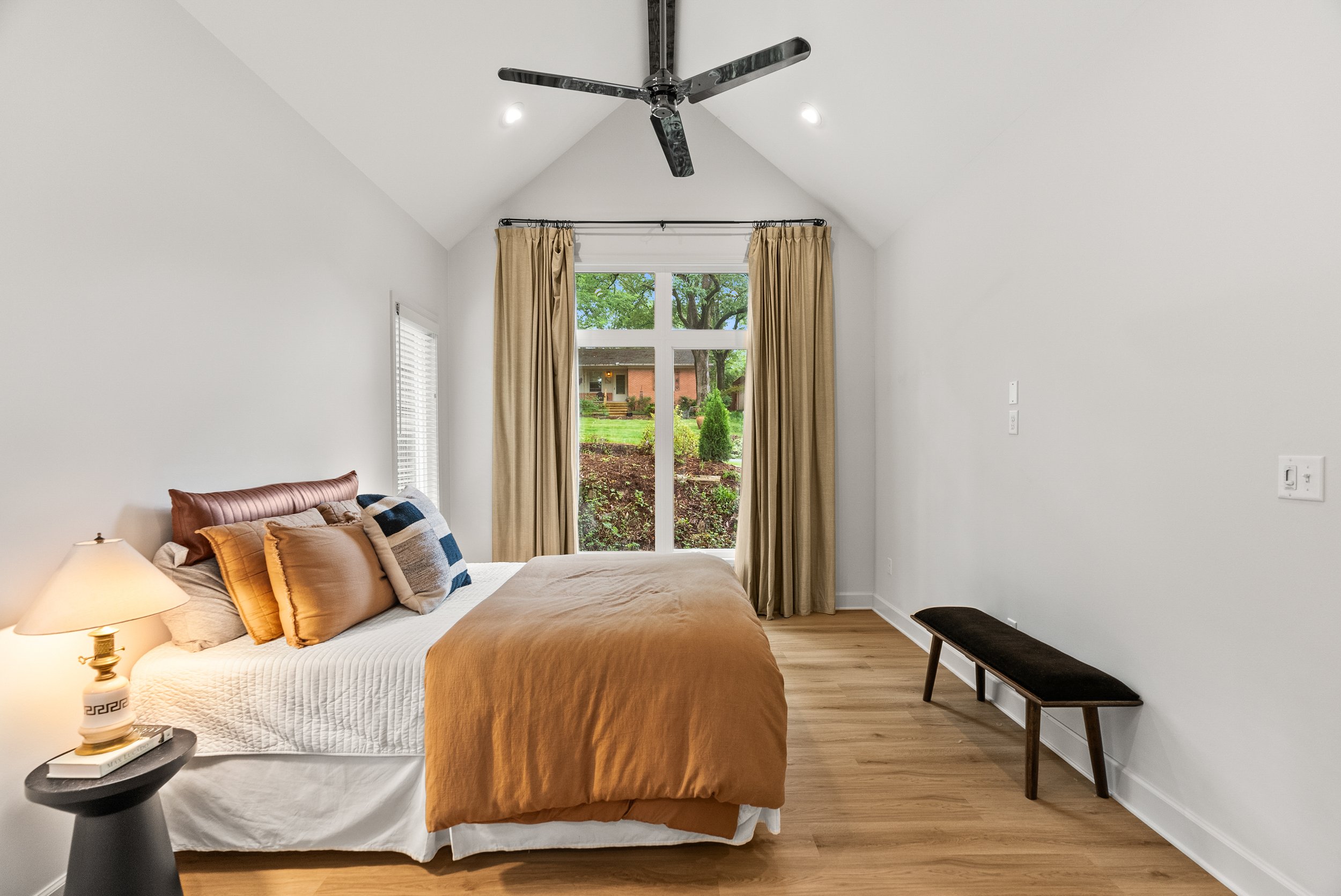 A bedroom with a large window and beige curtains, a bed with brown and beige pillows, a black side table with a lamp, and a bench against the wall, with a ceiling fan overhead and small recessed lights.