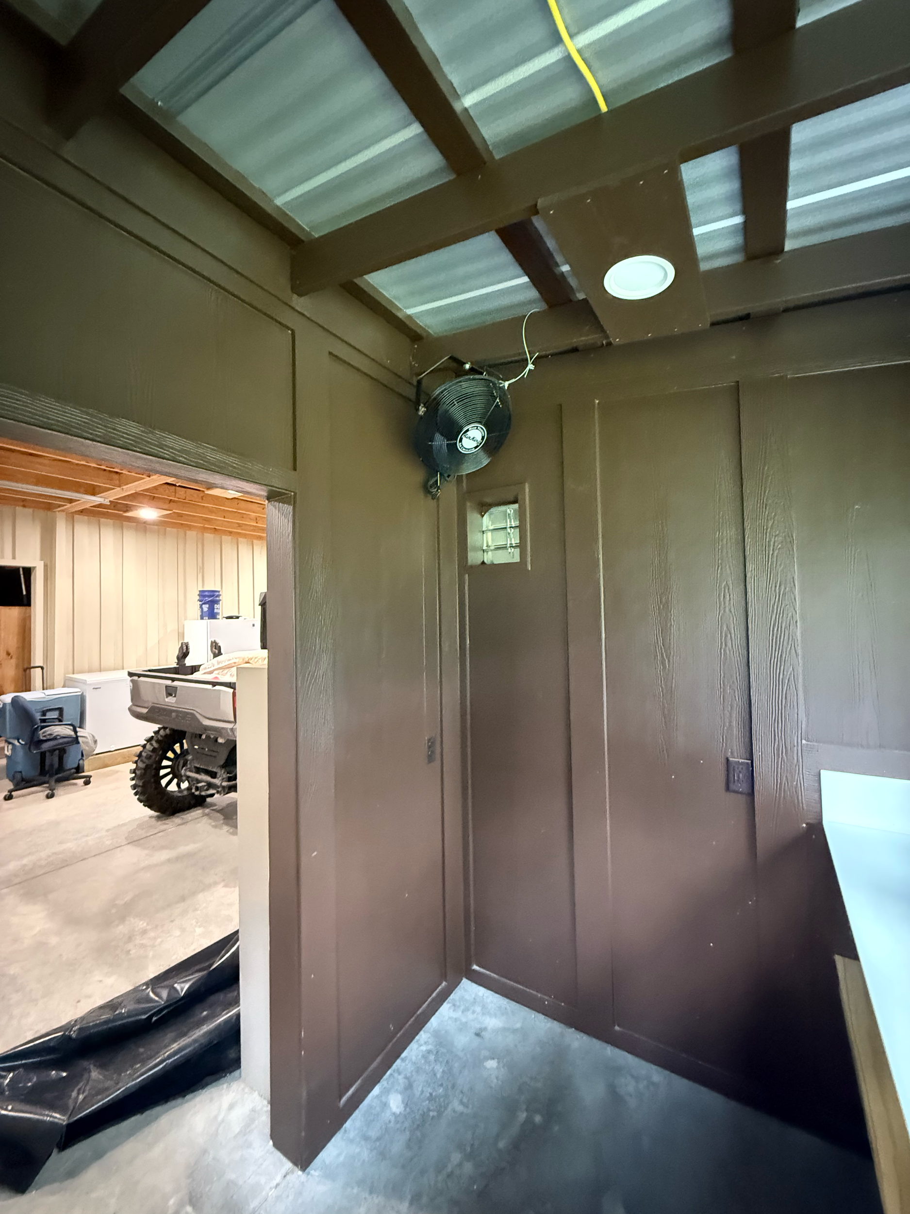 Photo of a corner in a room showing brown wooden ceiling beams, a wall-mounted black fan, and a doorway leading to a space with beige wooden walls and a concrete floor, with some stored items visible.