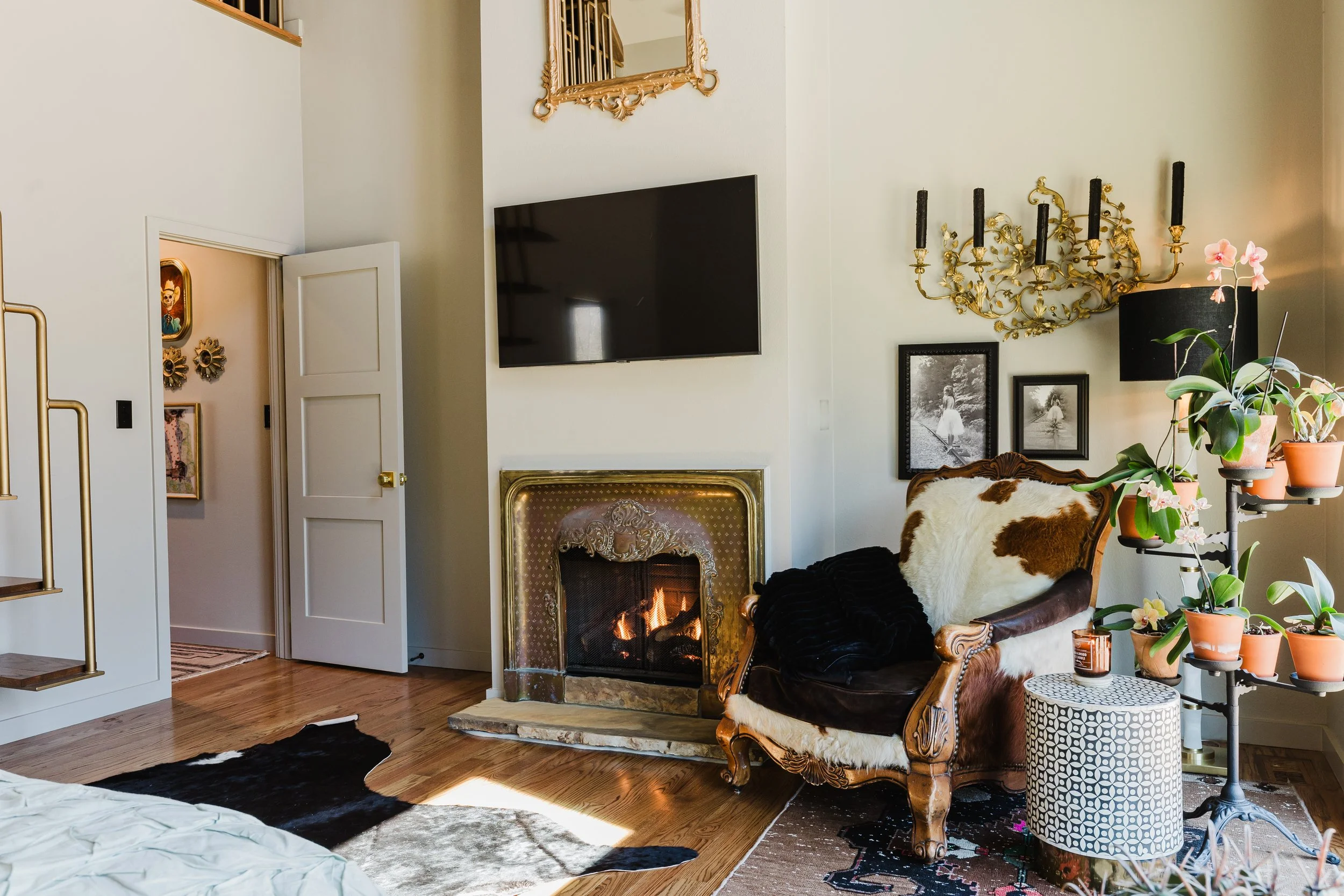 Living room with white walls, fireplace with ornate gold frame, vintage carved wooden armchair with animal print cushion, black throw blanket, potted plants on a black stand, black lampshade, gold and black wall sconce, wall frames, television mounte
