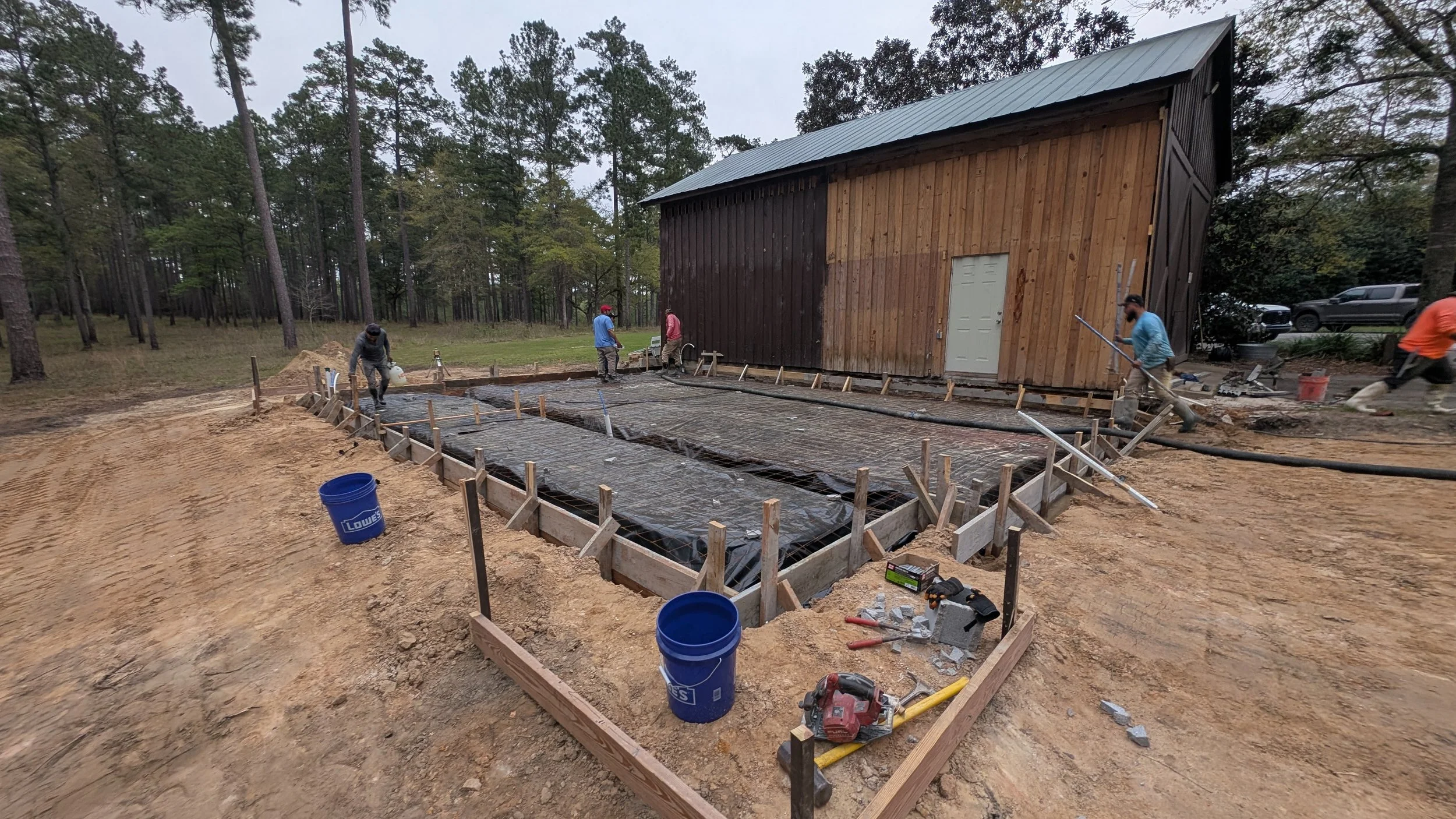 Construction workers pouring concrete on a foundation in front of a wooden shed surrounded by trees.