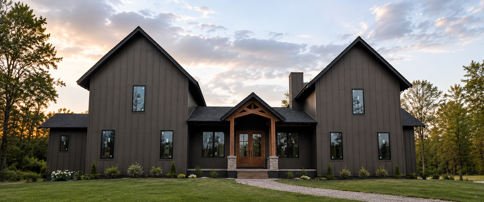 A modern house painted in dark gray with vertical siding, featuring two symmetrical gable roofs, a central entrance with wooden double doors, and large windows. The house is surrounded by a well-maintained lawn with small shrubs and trees, set against a backdrop of a partly cloudy sky during sunset.