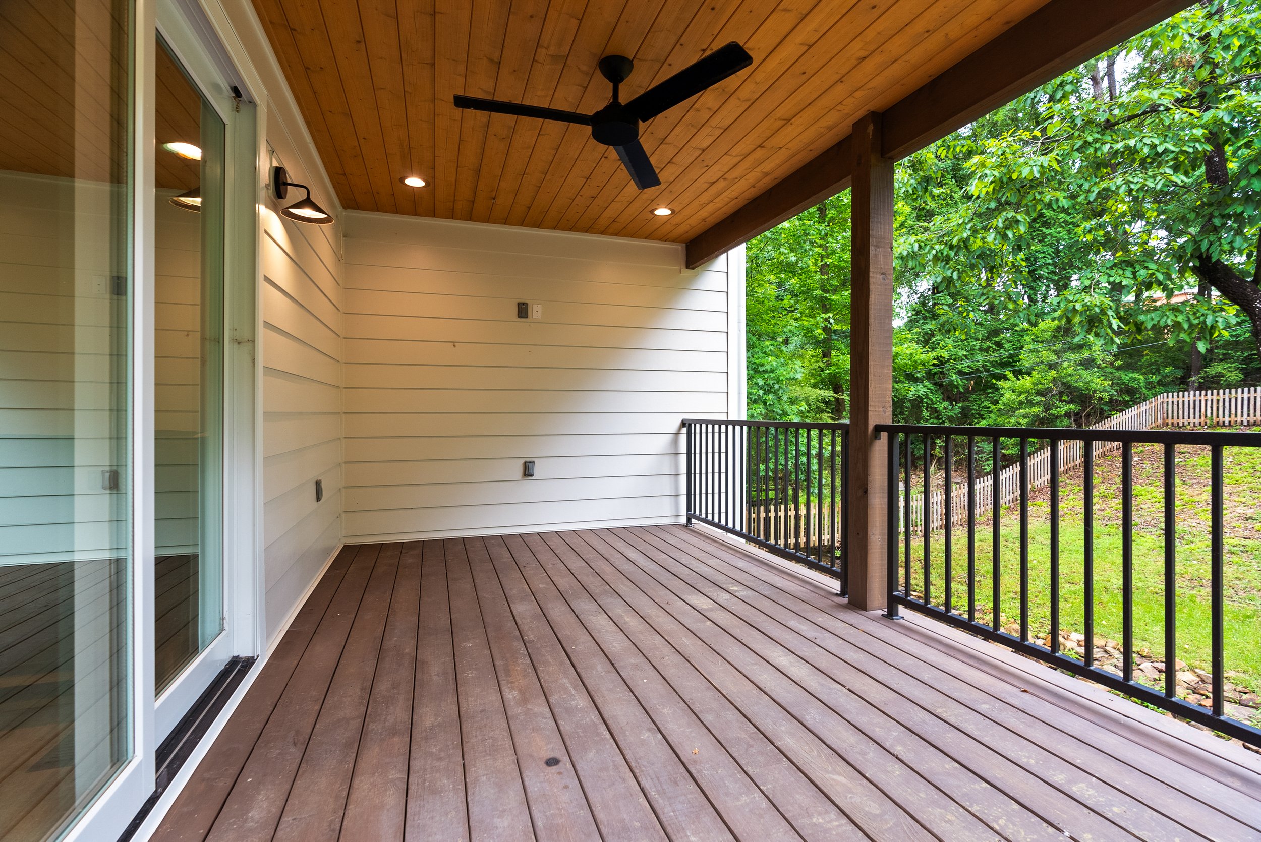 Image of a covered wooden porch with sliding glass door, black railing, ceiling fan, wood-paneled ceiling, and outdoor trees in the background.