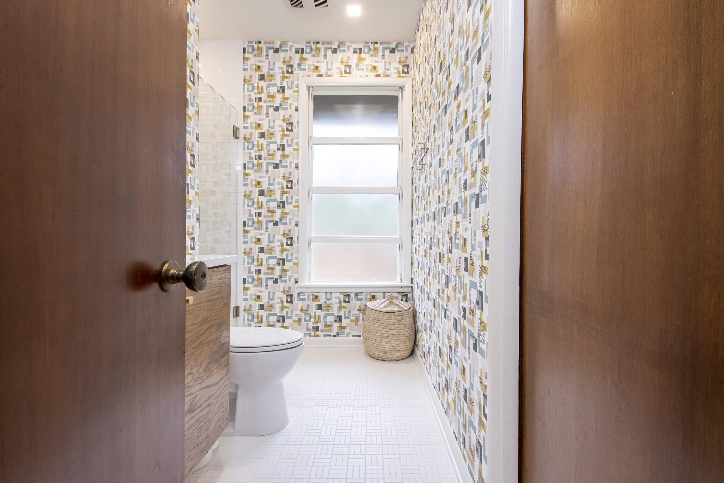 View of a brightly lit bathroom with geometric patterned wallpaper, a window, a wicker basket, and a toilet, seen from partially opened wooden door.