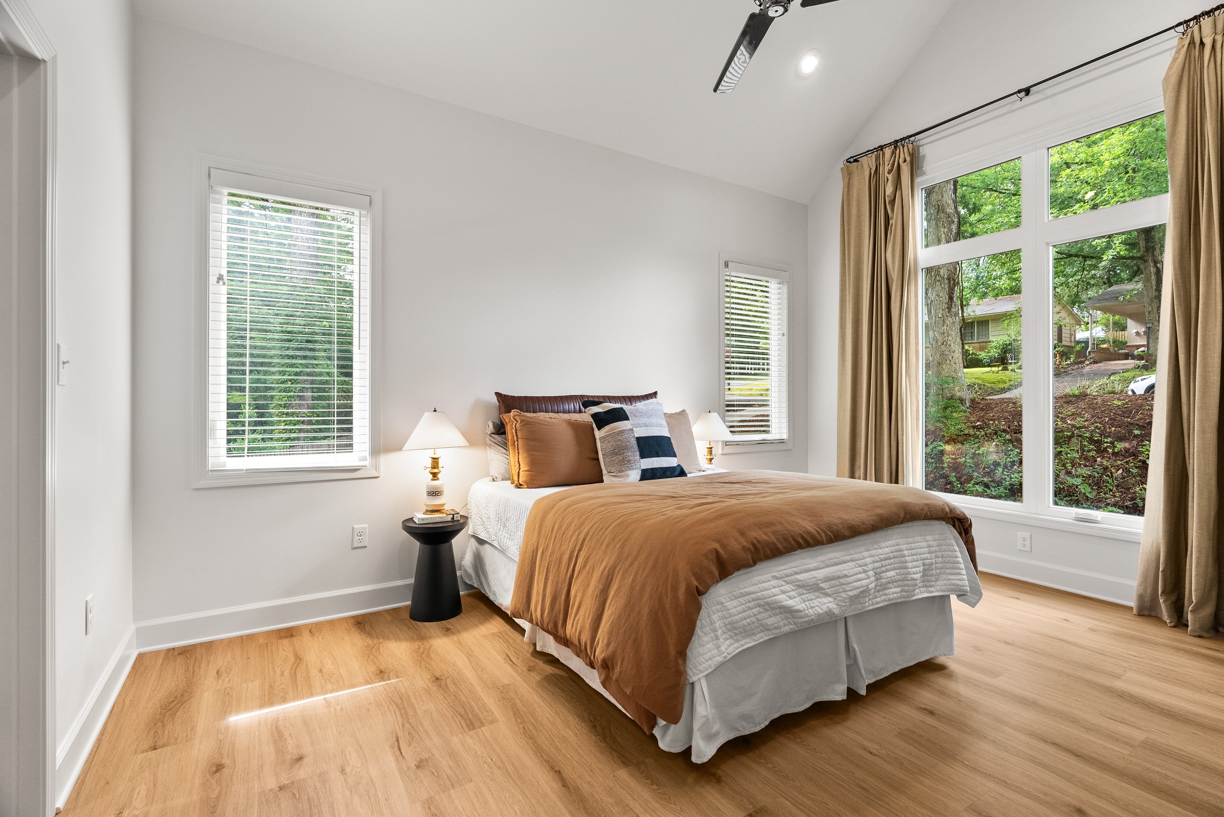 A bedroom with a large window showcasing trees outside, a bed with brown and black pillows, two white bedside lamps, and light-colored wooden flooring.