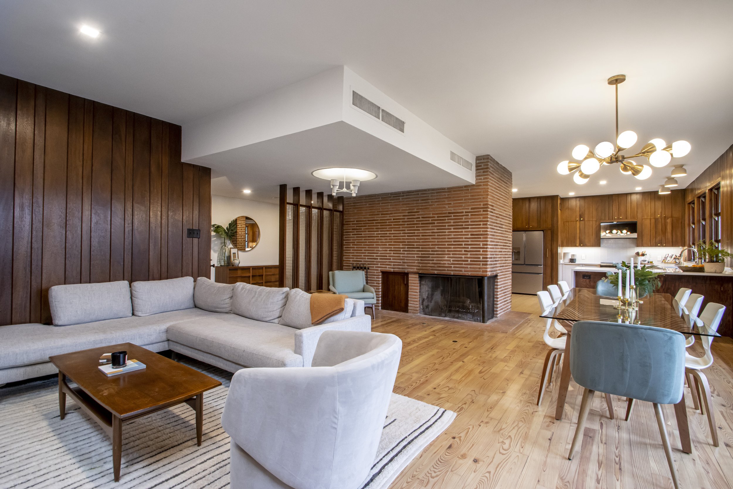 Open concept living and dining room with wood paneling, brick fireplace, modern lighting, and kitchen in the background.