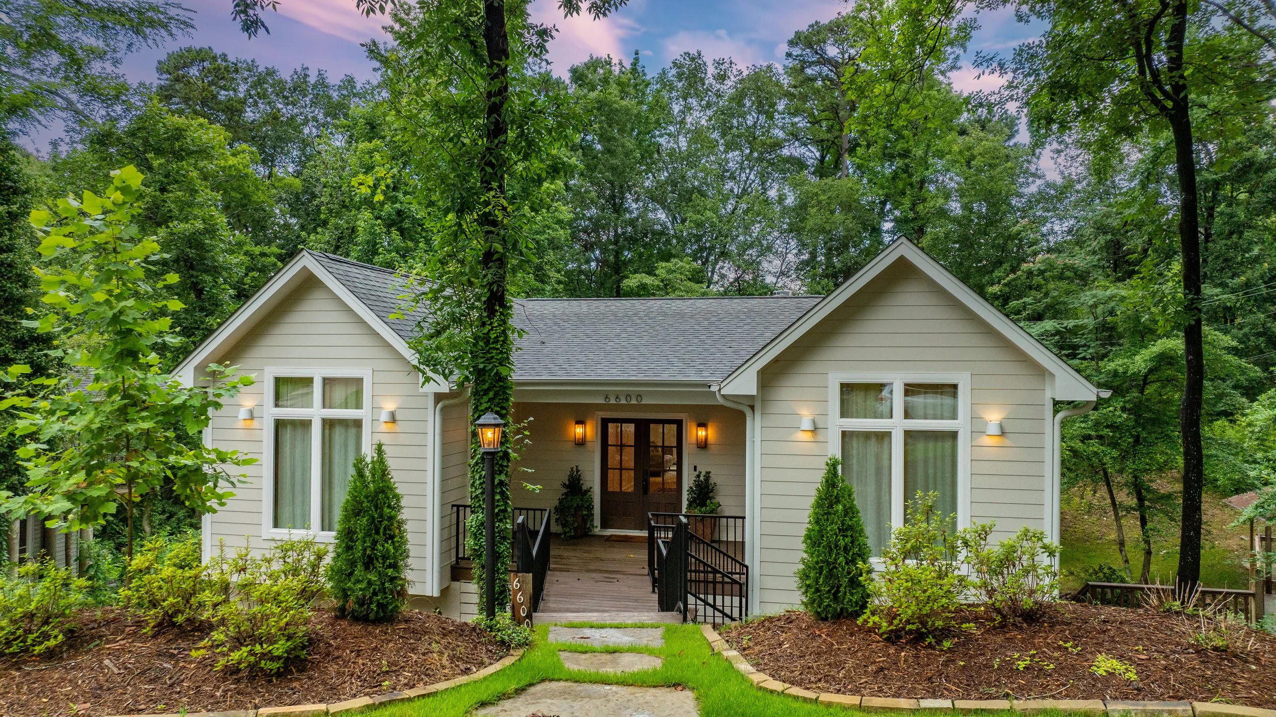 A cozy white house with a gray roof surrounded by lush green trees and a landscaped front yard with bushes and a walking path, illuminated by morning or evening light.