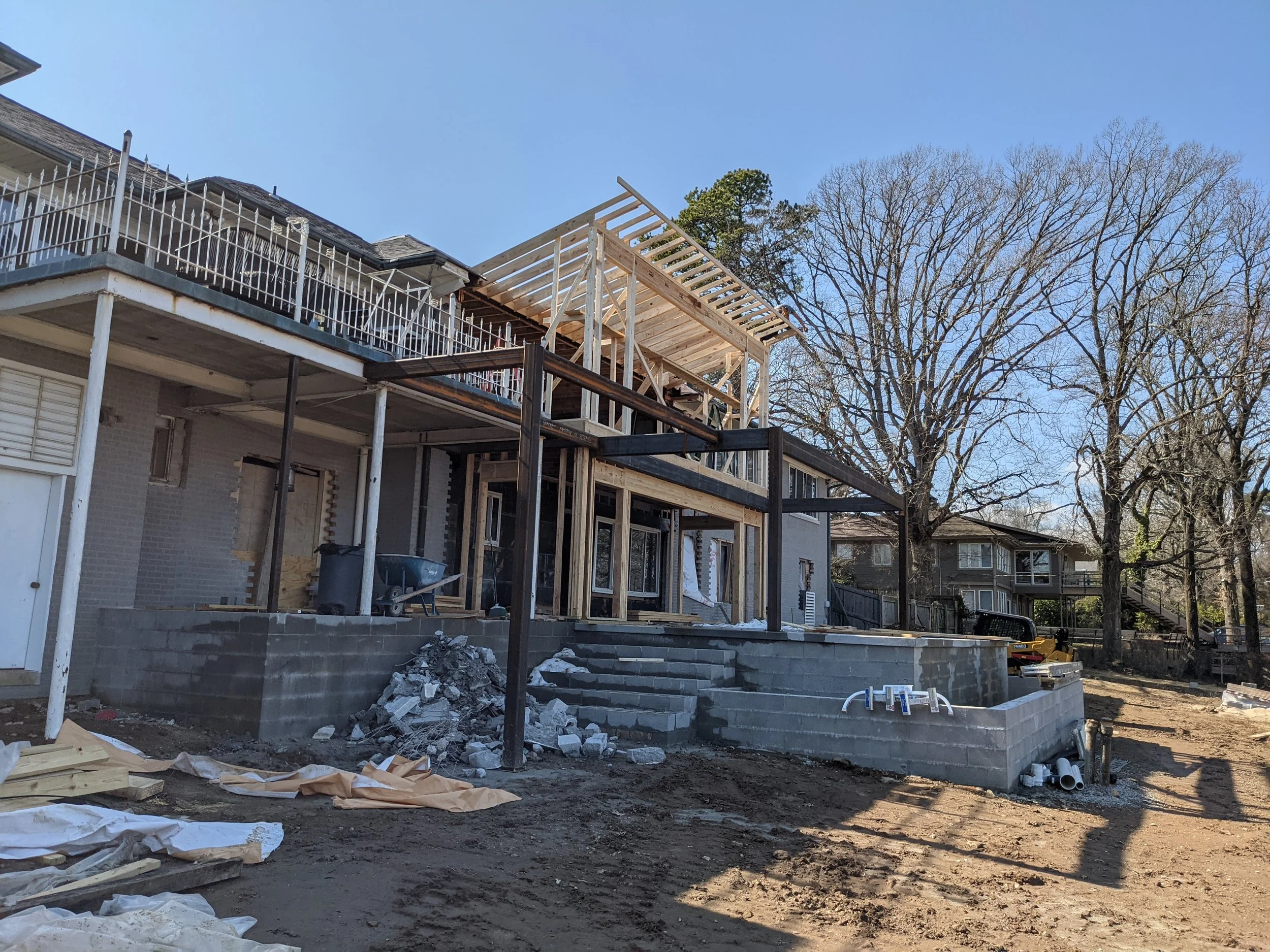 Construction site of a residential building with wooden framing on the upper level, gray brick walls, and a partially built staircase, with construction materials and tools scattered around, under a clear blue sky.