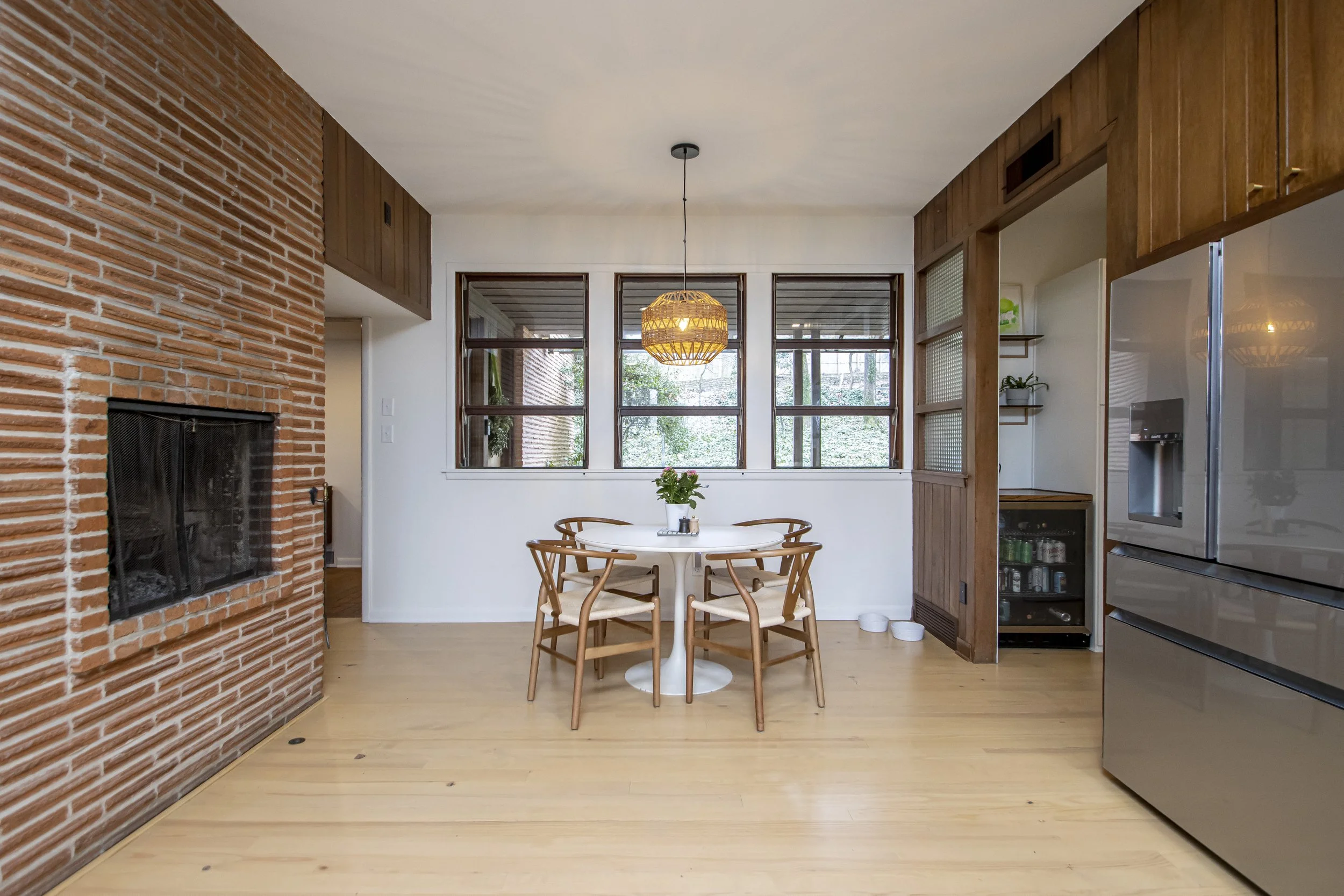 A cozy dining area in a home featuring a round white table with four wooden chairs, a window with a view of greenery, a pendant light hanging above the table, exposed brick wall, and wooden cabinetry.