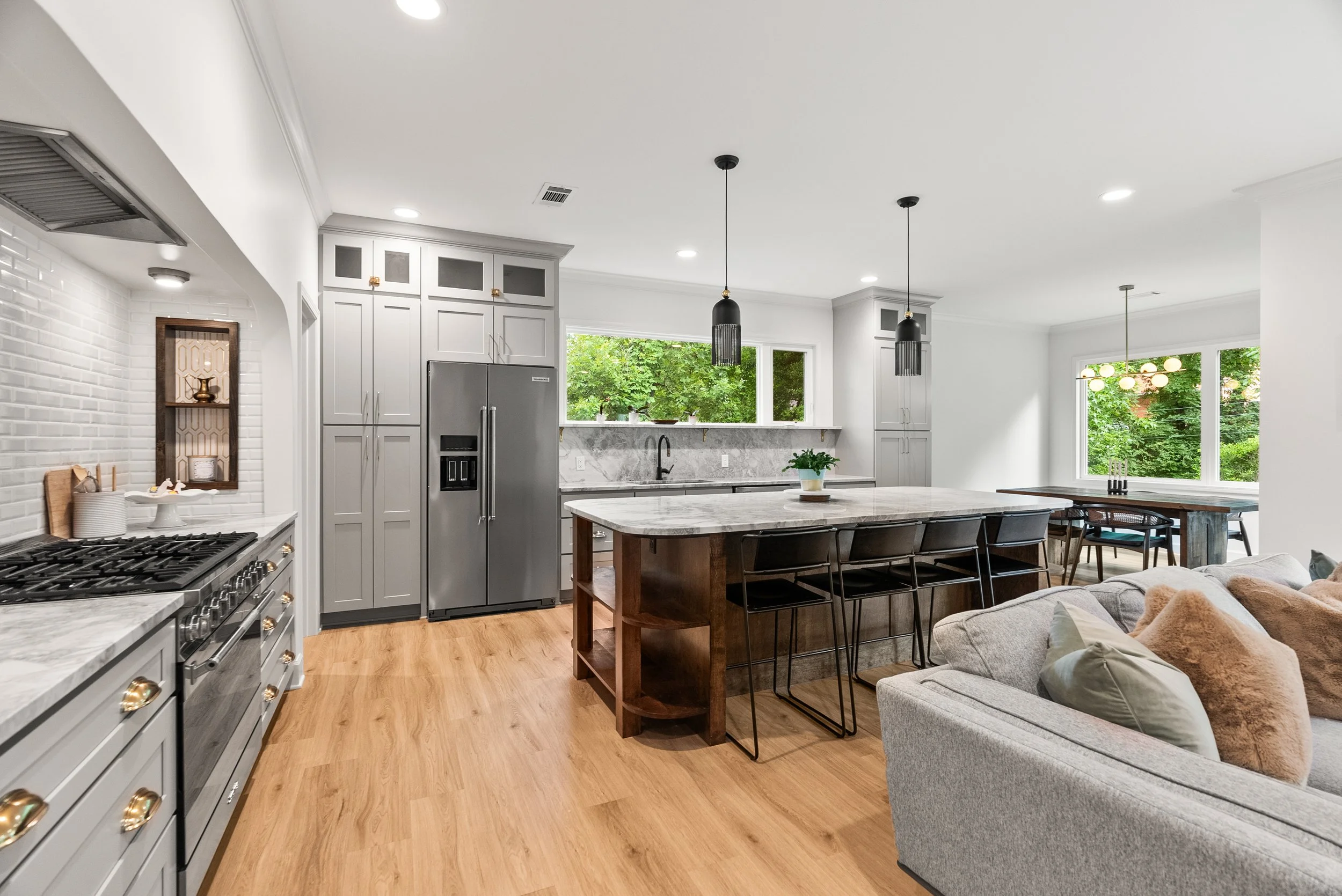 Modern open-concept kitchen and living area with large windows, gray cabinetry, marble countertops, and a gray sofa with pillows.