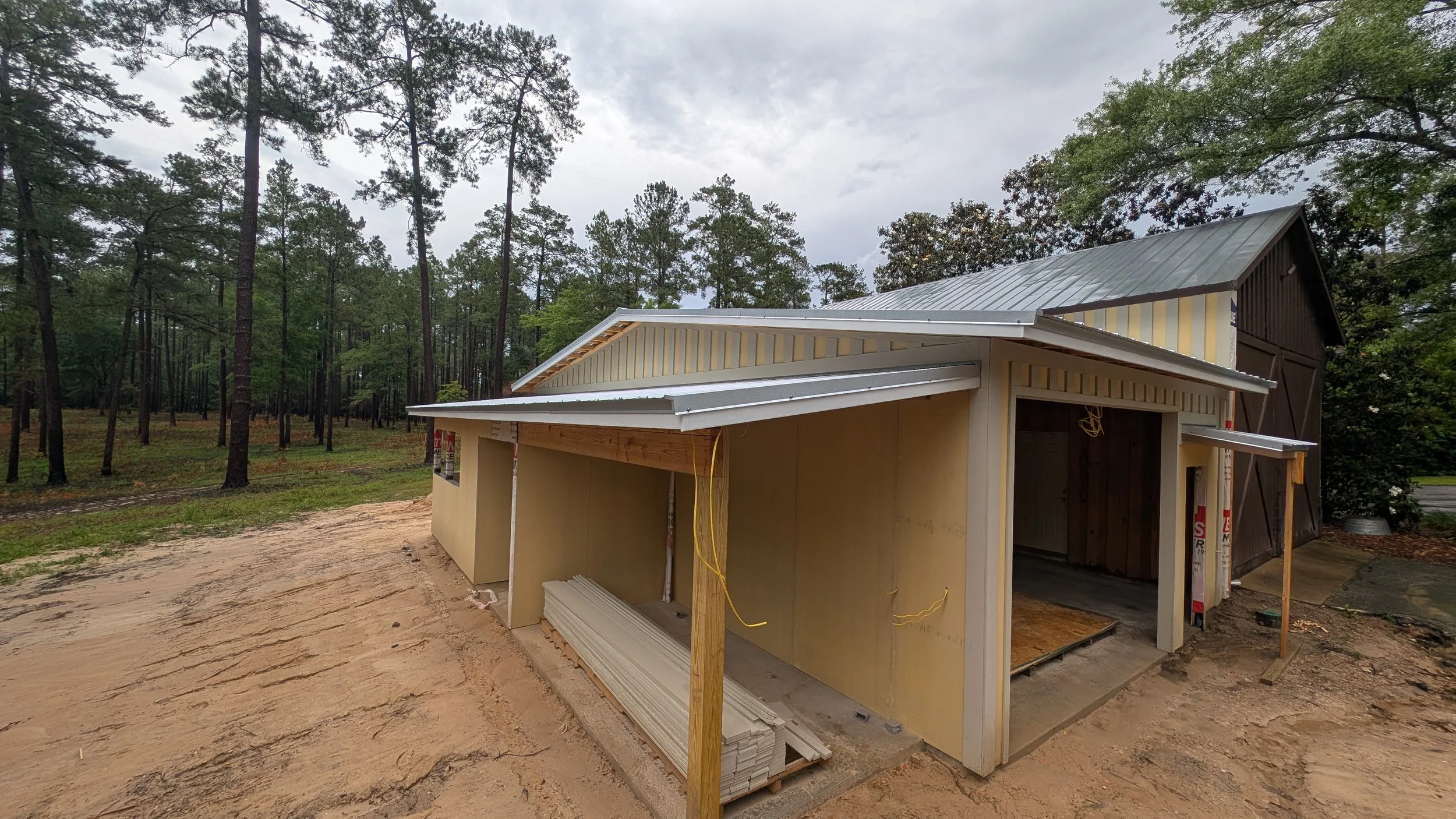 A building under construction in a wooded area with trees in the background. The structure has partially installed siding and roofing, with construction materials on the ground nearby.
