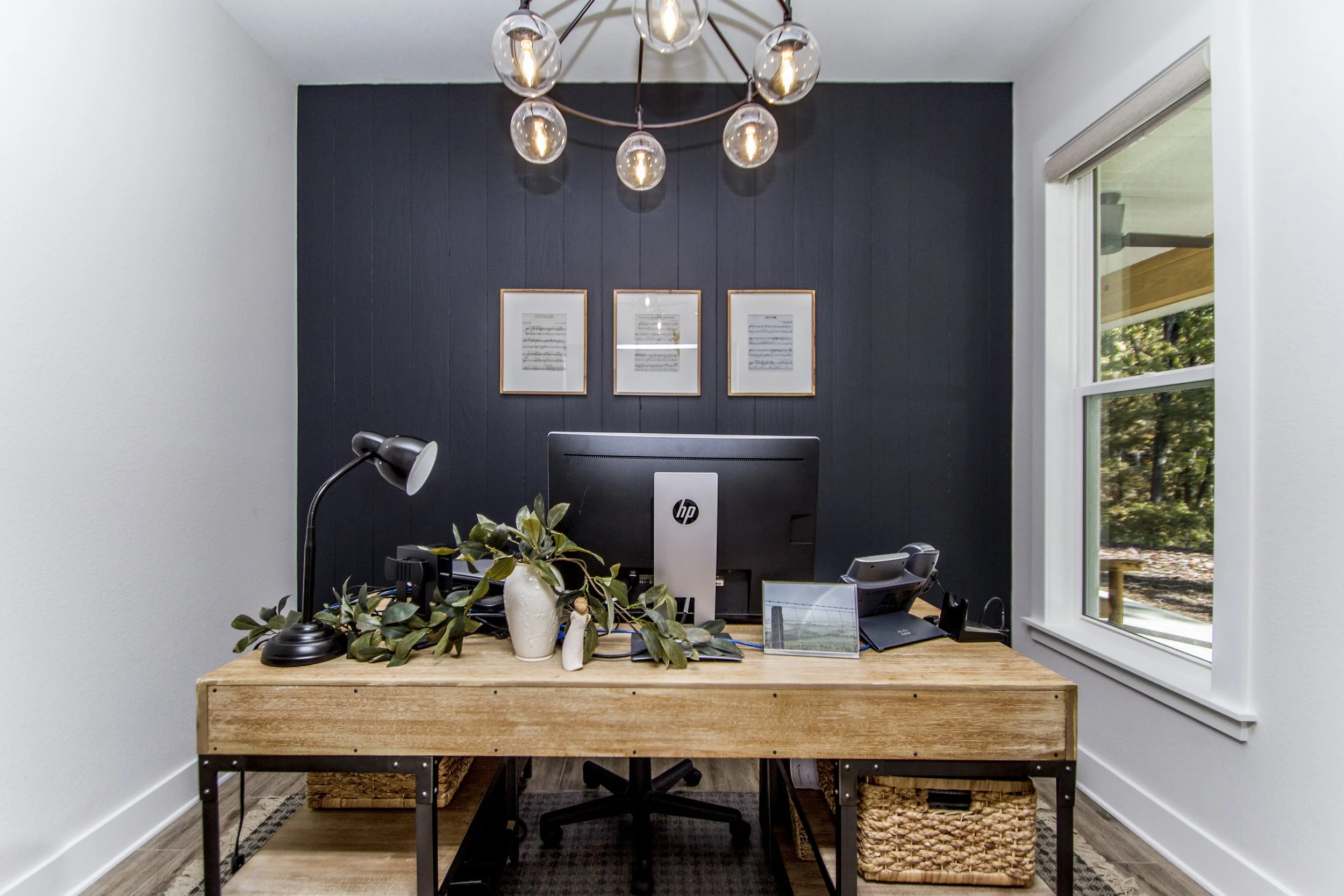 Office workspace with a wooden desk, black lamp, potted plant, computer monitor, framed pictures on a dark wall, window on the right, and a ceiling light fixture with exposed bulbs.