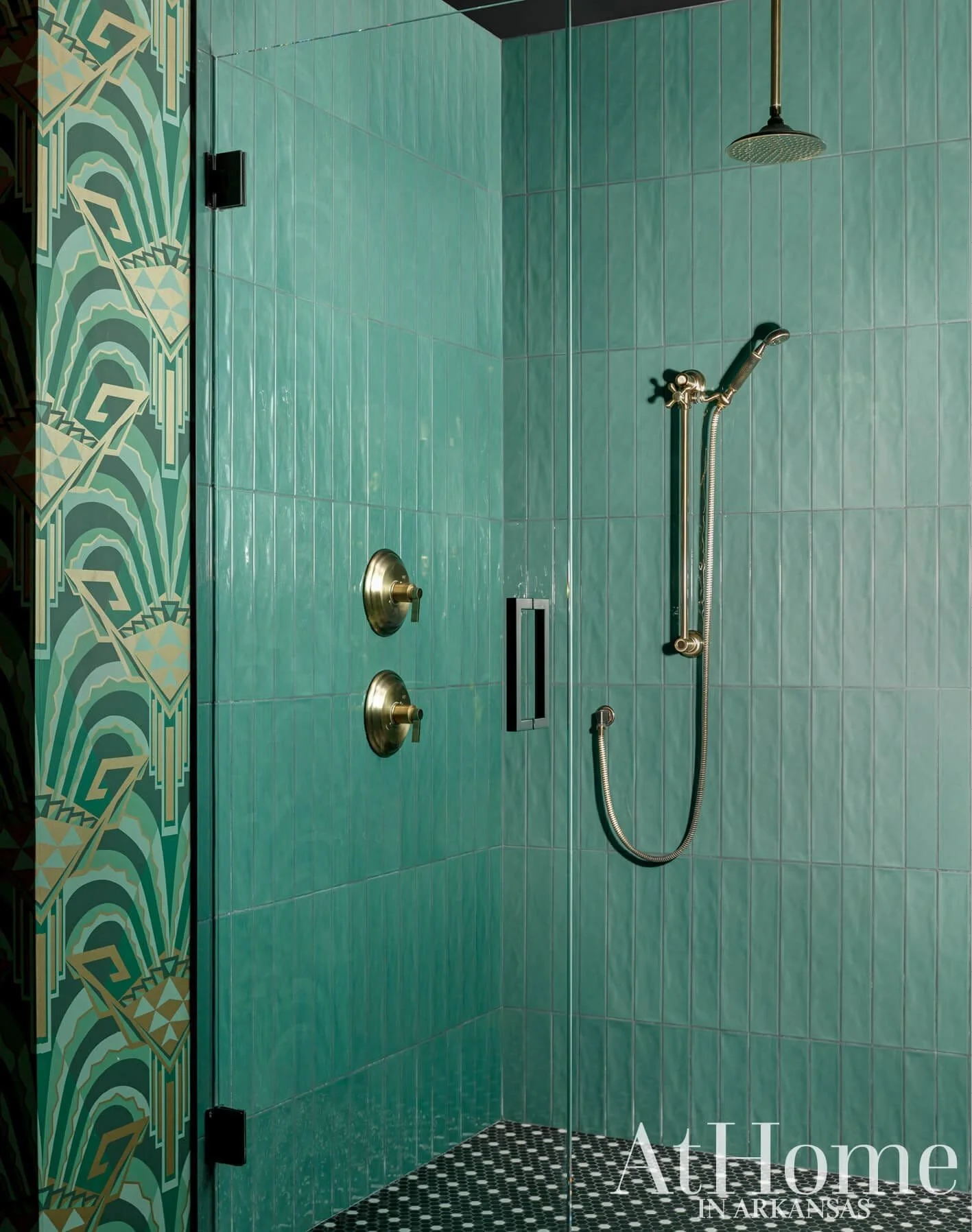 A walk-in shower with teal tiles on the walls, a rain showerhead, a handheld showerhead, two gold-colored fixtures, a glass door handle, and patterned black and white floor tiles. The bathroom wall features decorative geometric patterned tile.