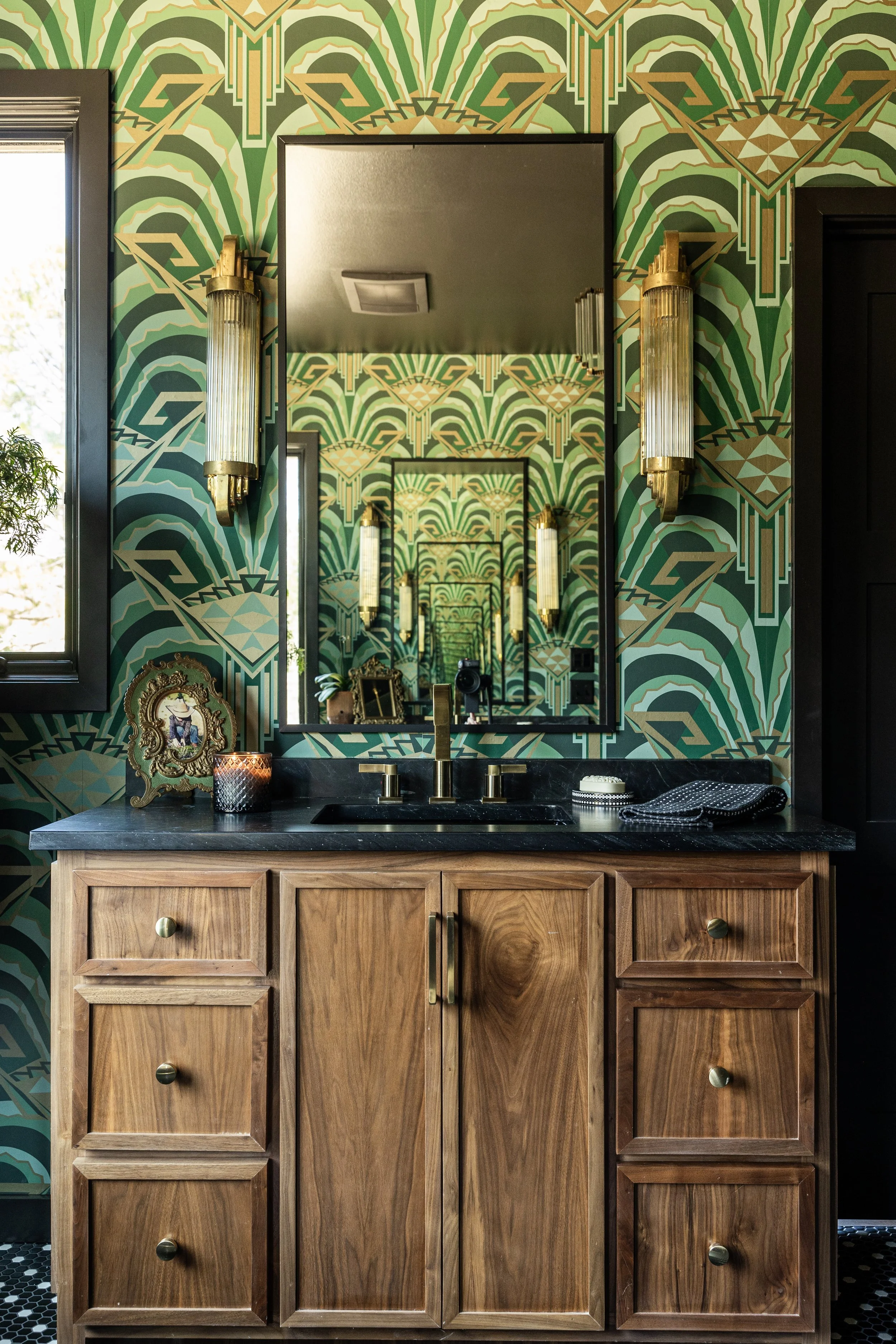 Bathroom sink vanity area with a wooden cabinet, black countertop, and a large mirror reflecting patterned green and gold wallpaper. Wall-mounted gold fixtures with glass covers flank the mirror, and decorative items including a framed picture, candl