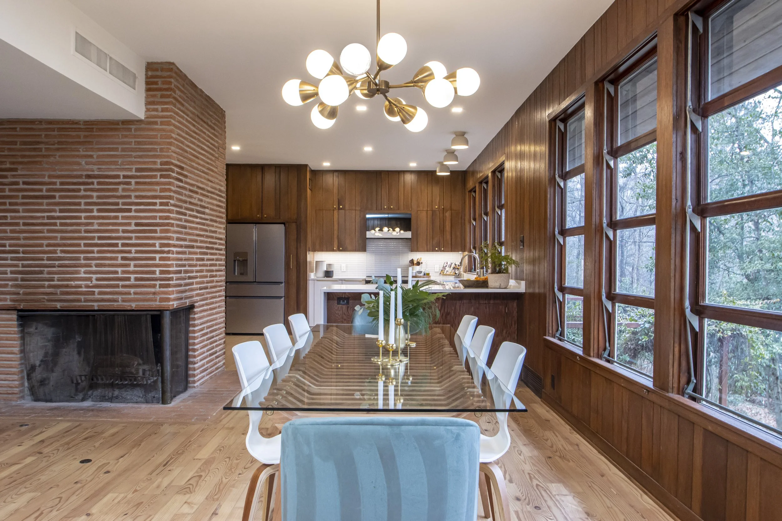 Modern dining room with large wooden table, white chairs, brick fireplace, wood-paneled walls, large windows with views of trees, and contemporary chandelier lighting.