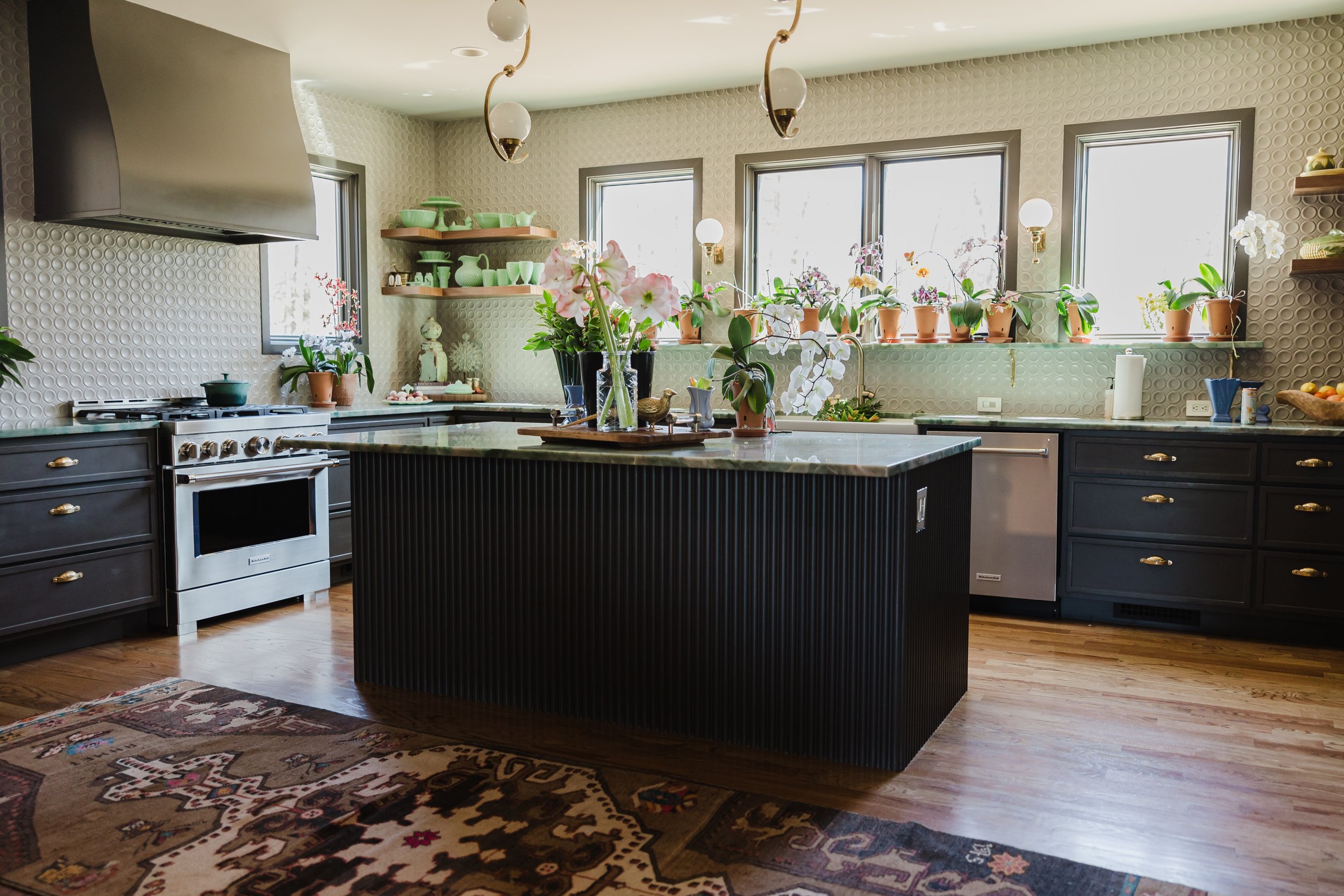 A modern kitchen with a central island, dark cabinets, and a white stove. The kitchen features multiple windows with potted orchids on the windowsills, and the walls have a textured geometric pattern. There are various decorative items and a colorful