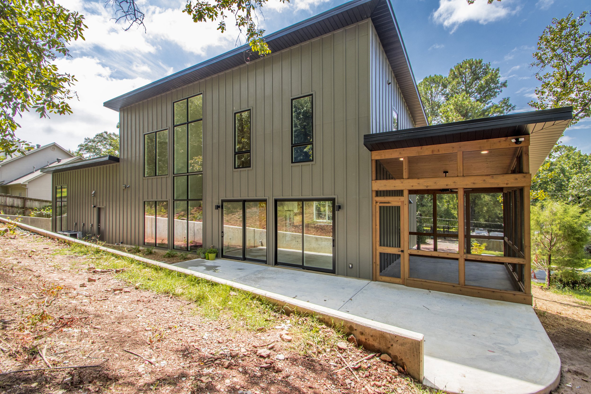 Modern two-story house with vertical metal siding, large windows, and a covered wooden porch in a backyard with trees and a concrete patio.