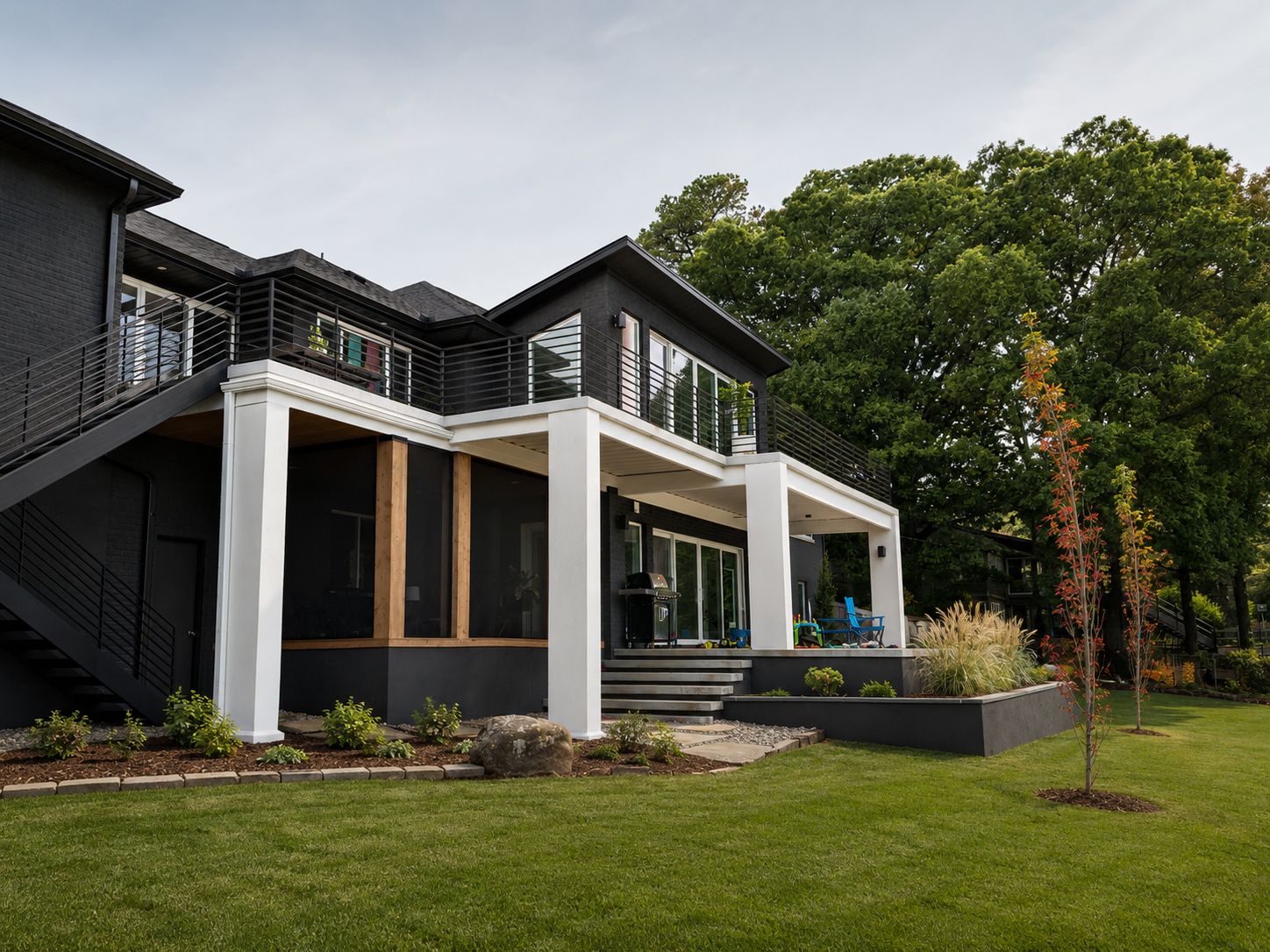 Modern black and white house with a large balcony, outdoor stairs, and a backyard with manicured grass, trees, and plants.