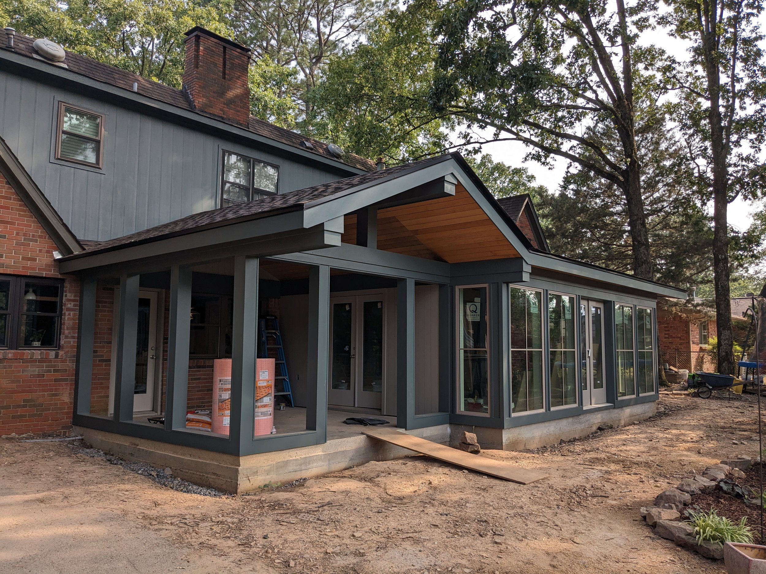 Rear view of a house under construction with a large covered patio area, new sliding glass doors, and a mix of brick and siding exterior. There is a chimney and stacks of building materials visible, with trees in the background.