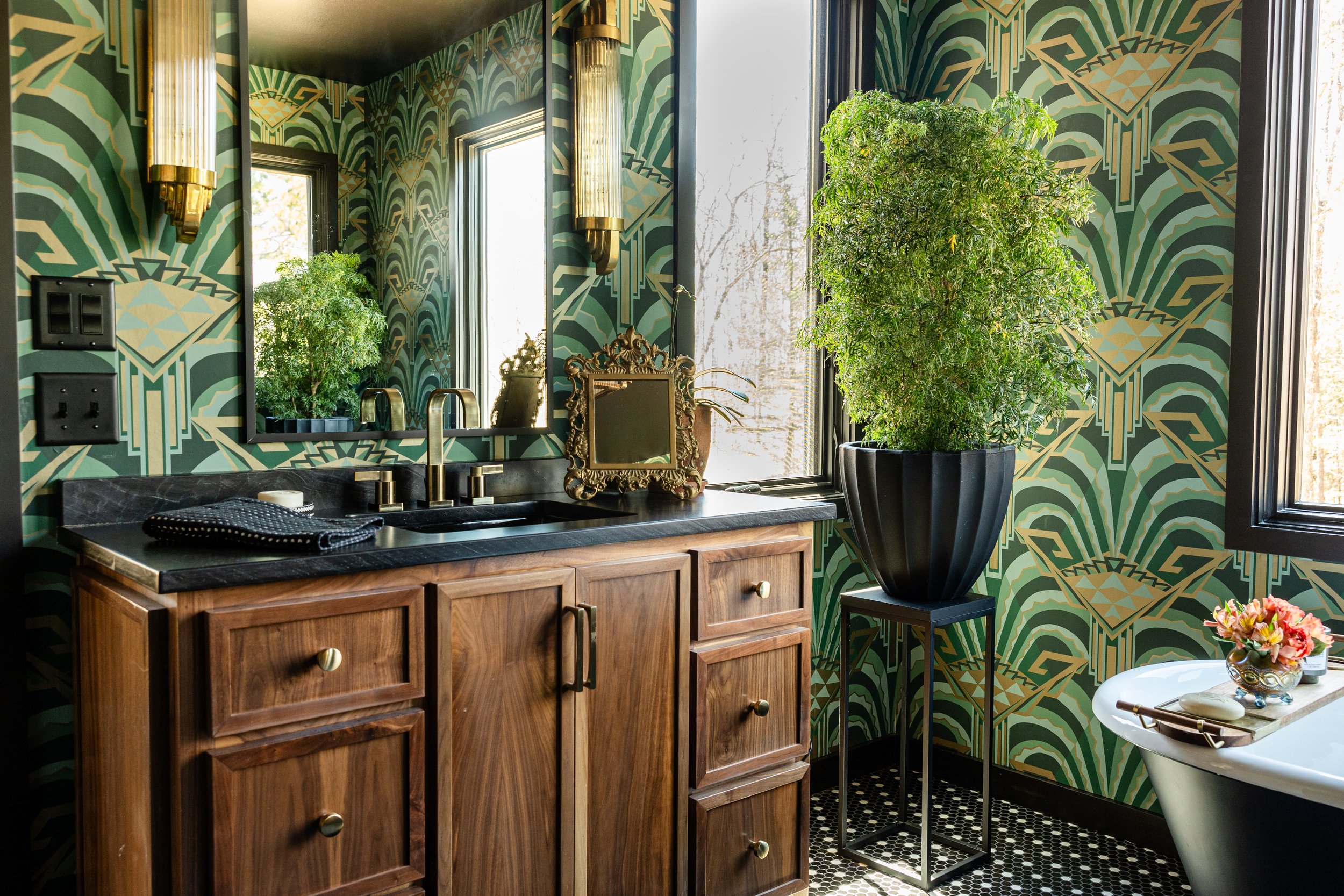 A bathroom with geometric green and gold wallpaper, a large potted plant, a wooden cabinet with a black countertop, a mirror, gold wall sconce lights, and decorative items on the counter.
